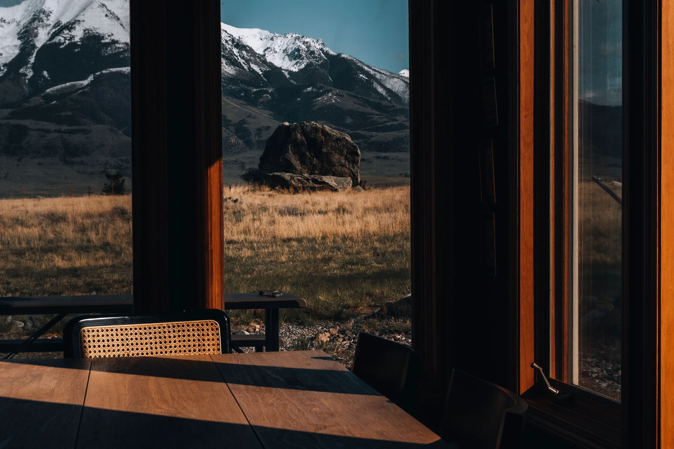 View of snow-capped Absaroka mountains through the dining room window with dark frames, capturing a grassy field and large rock outside.