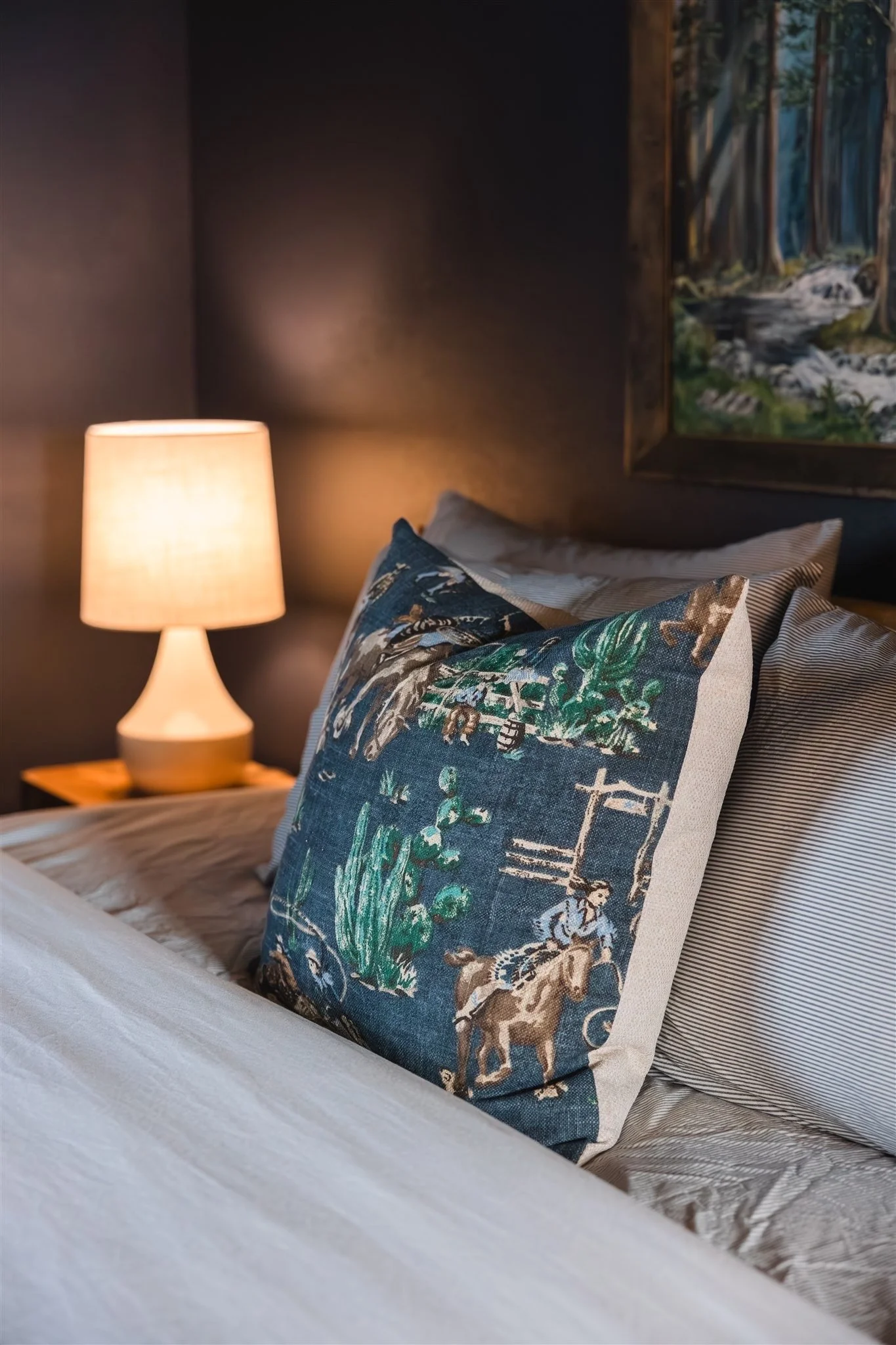 A close-up of a bed with decorative pillows, a nightstand with a lit lamp, and a landscape painting of a forest with a creek on the wall.