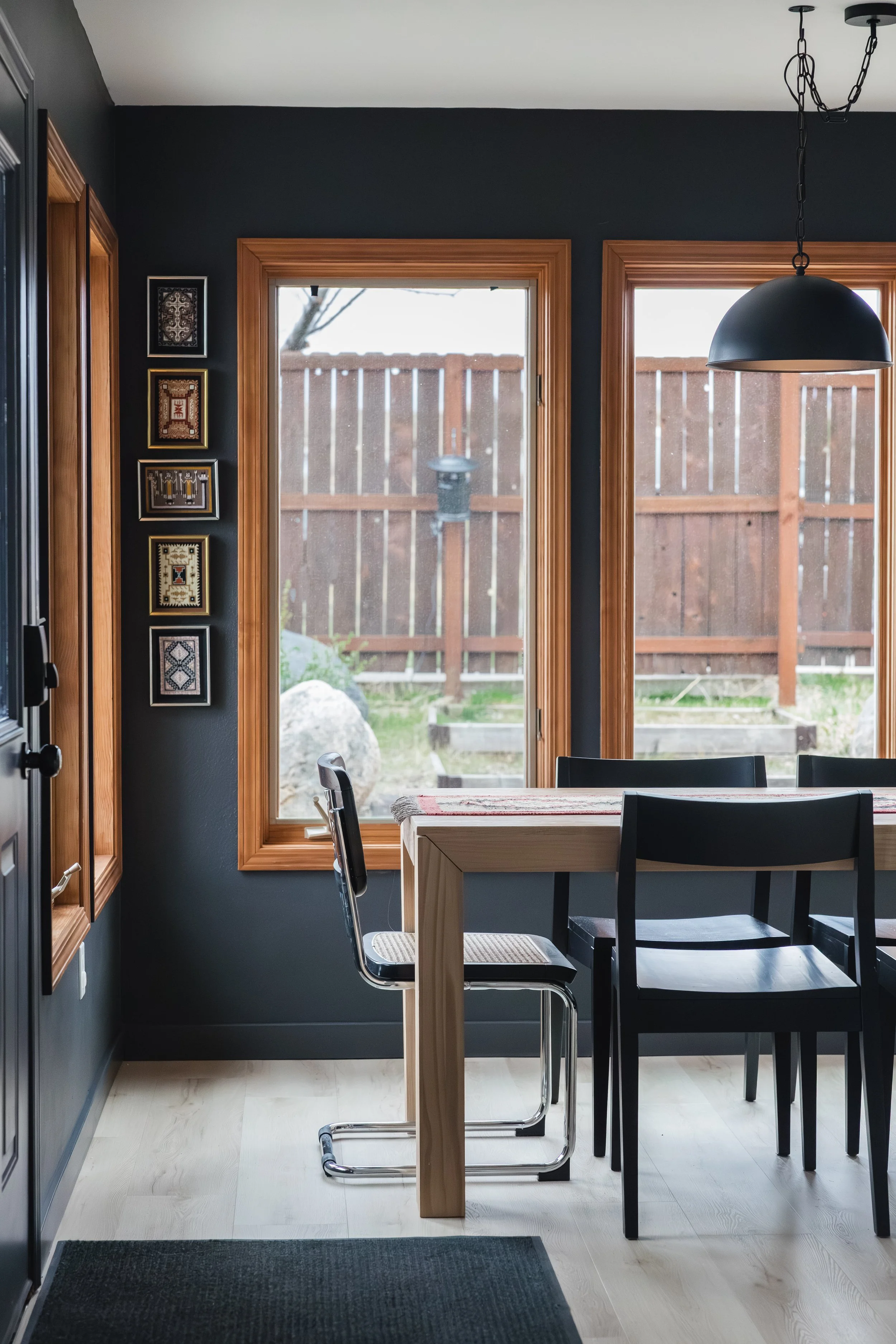 A dining area with large windows, a wooden table, black chairs, and a black pendant light. There are framed artworks on a dark-colored wall, and outside the windows, a wooden fence and some rocks are visible.