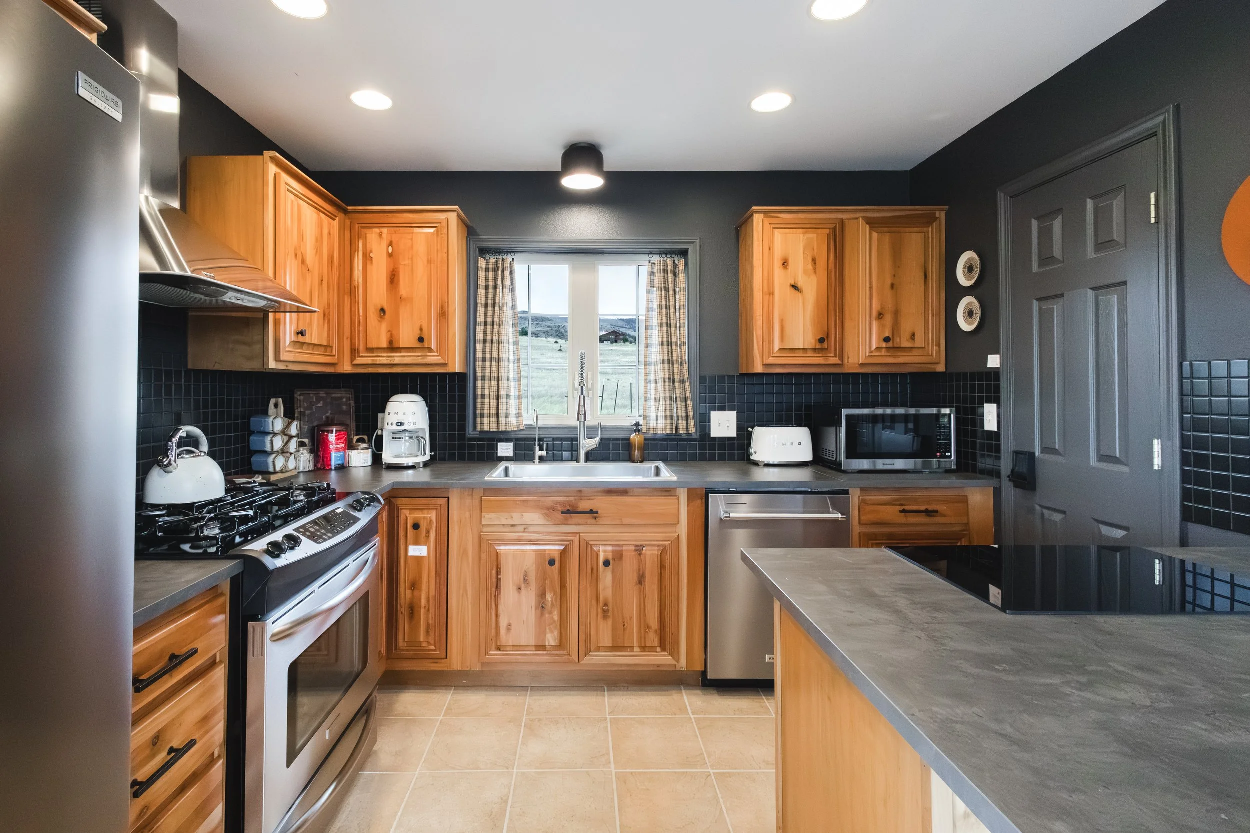 A modern kitchen with wooden cabinets, black backsplash tiles, a stainless steel stove and microwave, a white toaster, a coffee maker, and a window with plaid curtains overlooking open land.