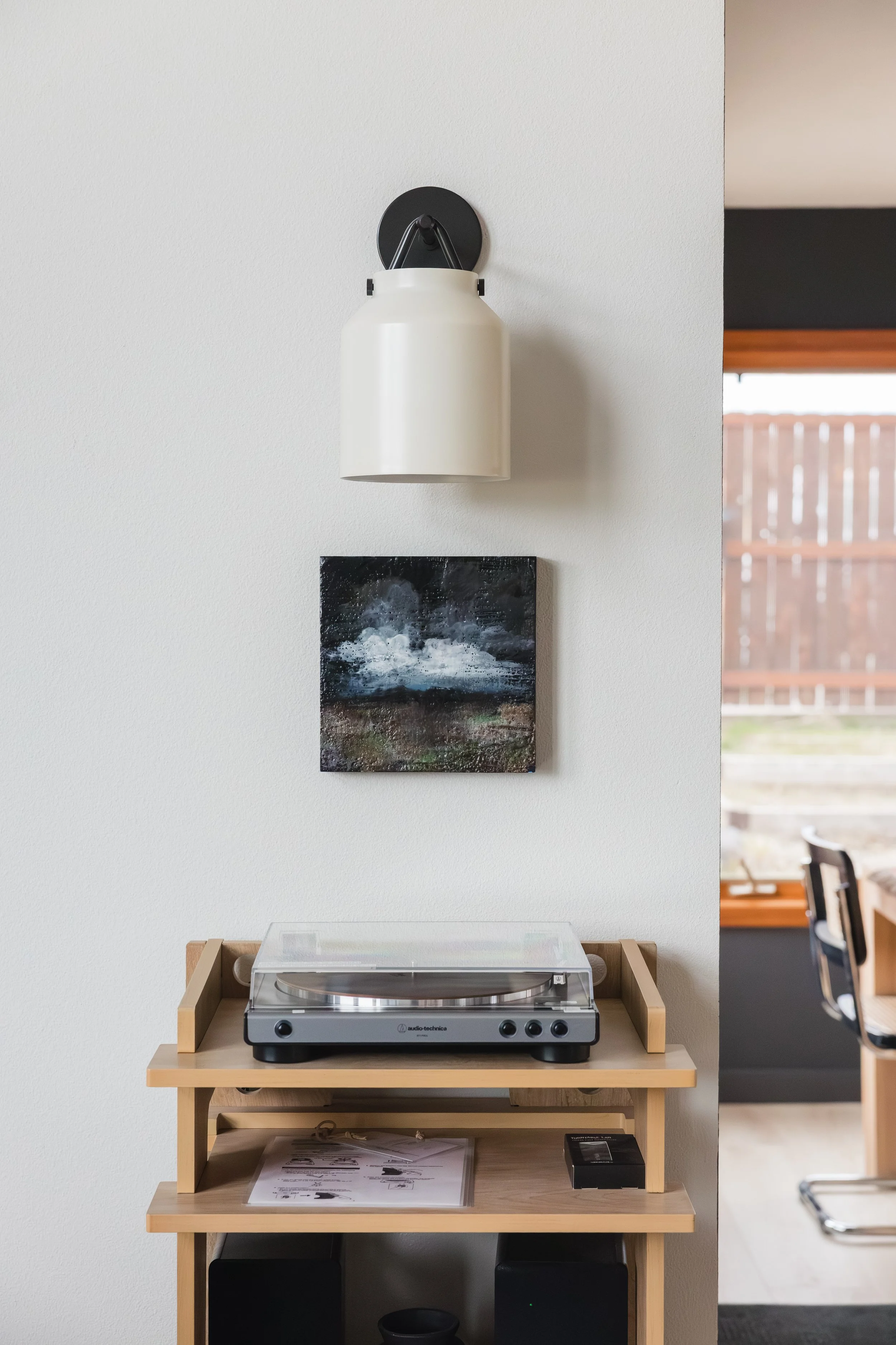 A wooden table with a turntable in a wooden frame on top, positioned against a white wall. Above the table, a white wall lamp is mounted, and below it, an abstract landscape painting. To the right, a small part of a chair and a window with wooden bli