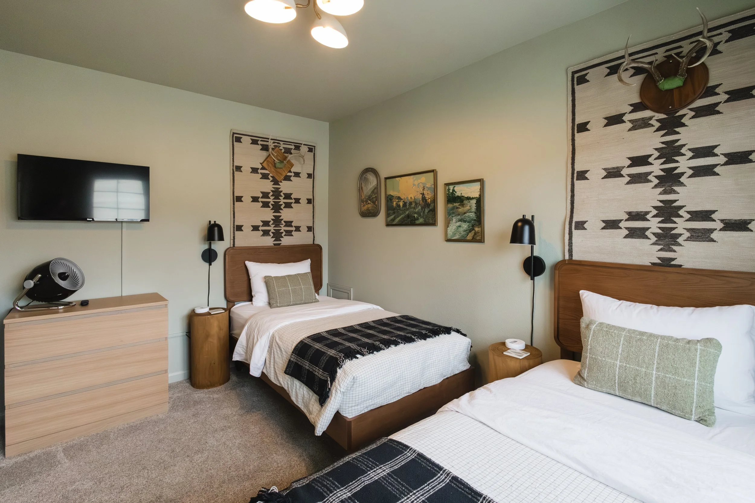 A cozy bedroom with two beds, wooden headboards, black and white patterned throws, and pillows. A dresser with a fan, black wall sconces, a mounted TV, and three landscape paintings on the wall. Decor includes tribal-style woven wall hangings and ant