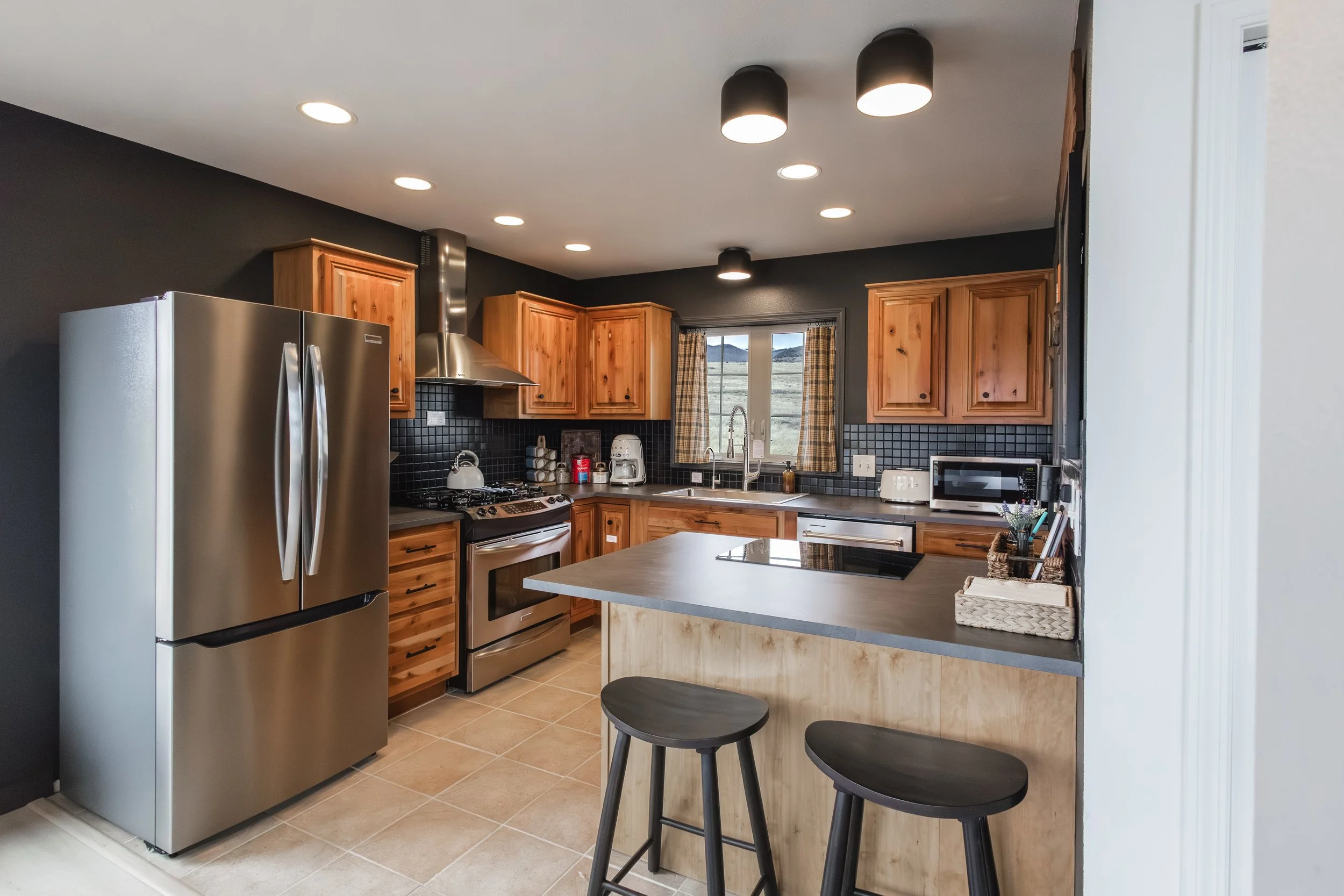 Kitchen with stainless steel refrigerator, wooden cabinets, black tiled backsplash, window with plaid curtains, and island with two black stools.
