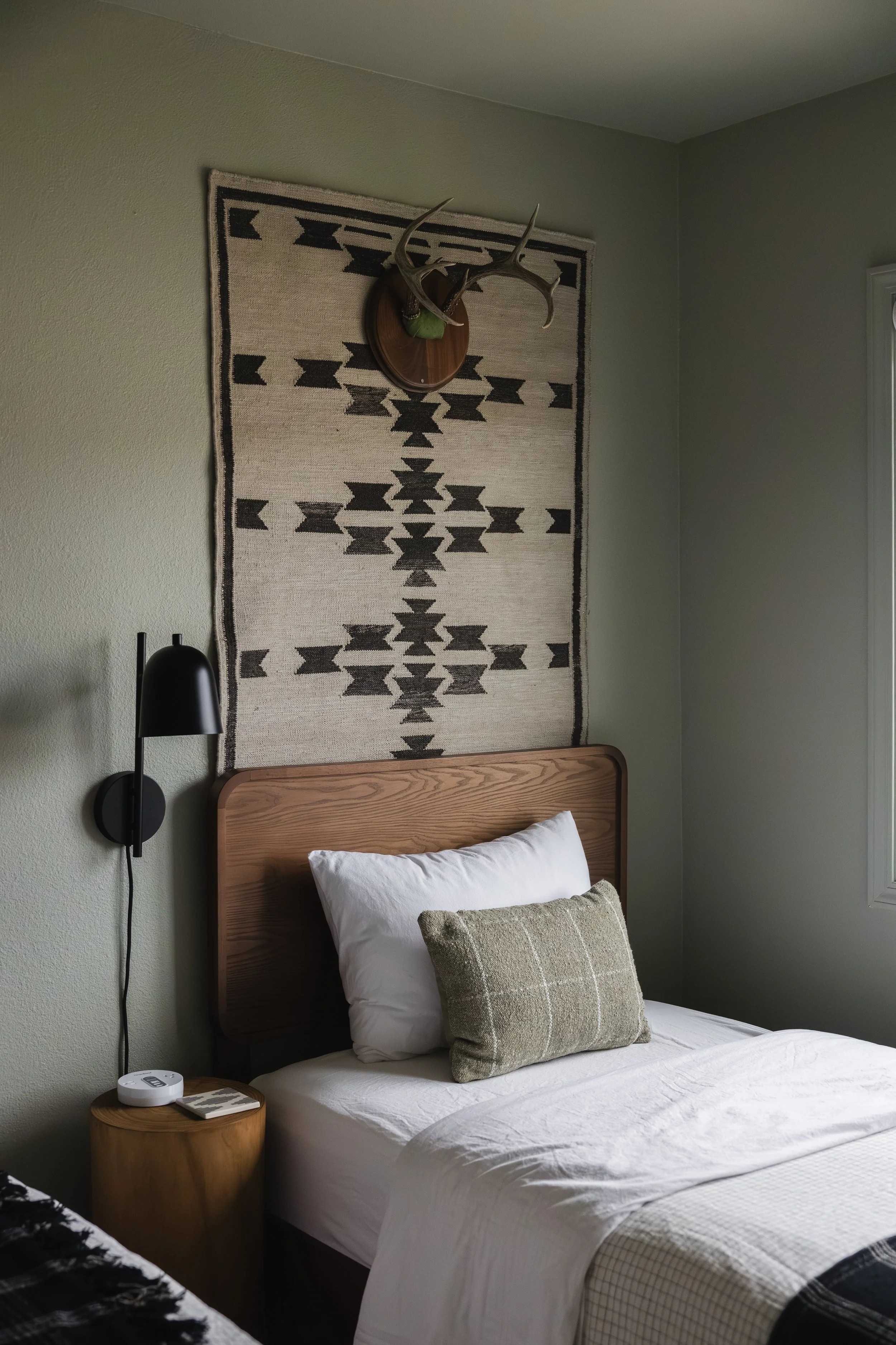 A bedroom with a wooden bed frame, white bedding, a beige pillow, and a gray pillow. A black wall lamp is mounted on the wall next to the bed. A nightstand beside the bed holds a white clock and a remote control. Behind the bed, there is a decorative