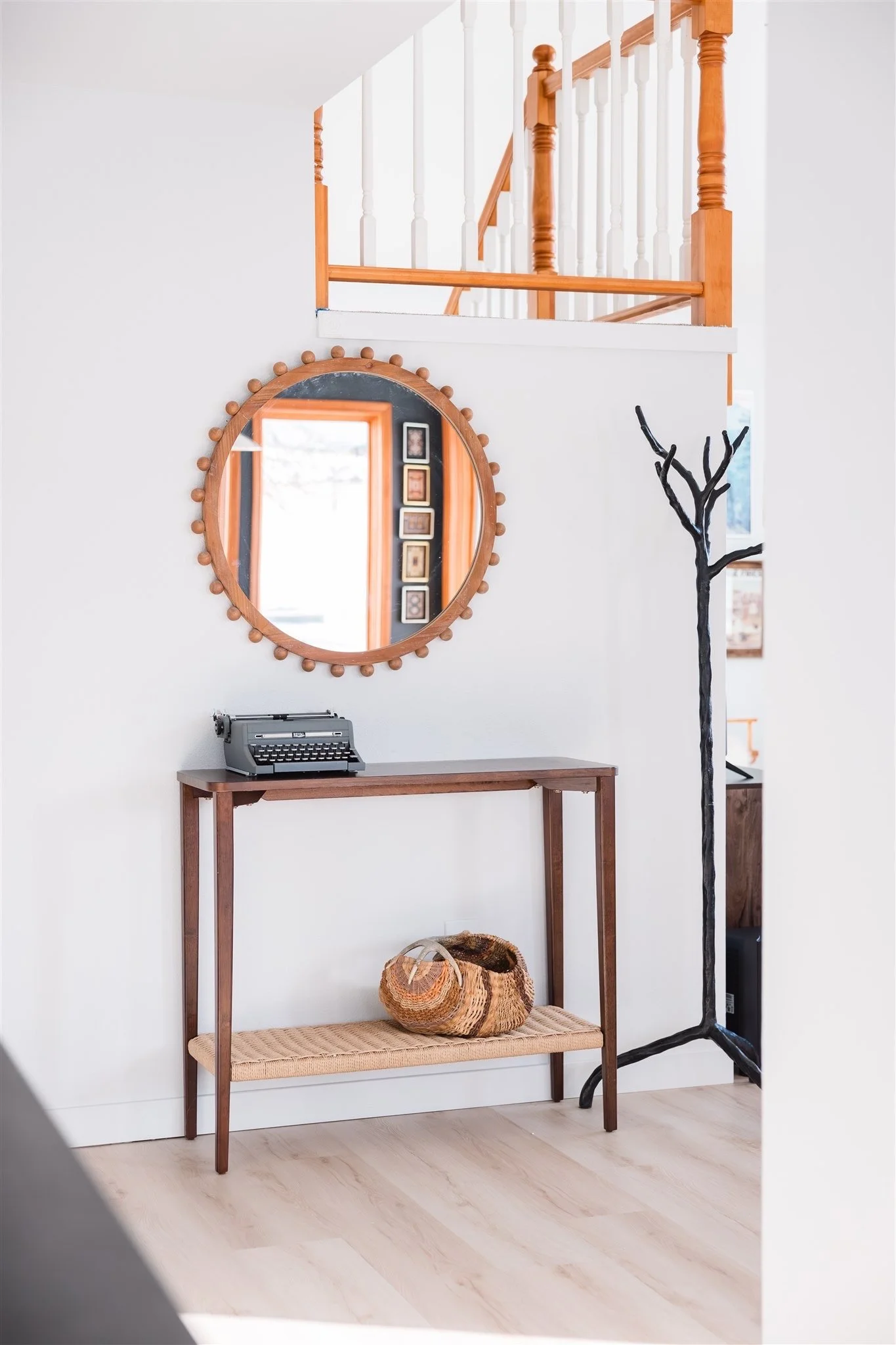 Interior of a home with a white wall, a wooden table, a woven basket on a lower shelf, a round mirror with wooden beads, a vintage typewriter, a black painted bare tree sculpture, and an upstairs landing with wooden railing.