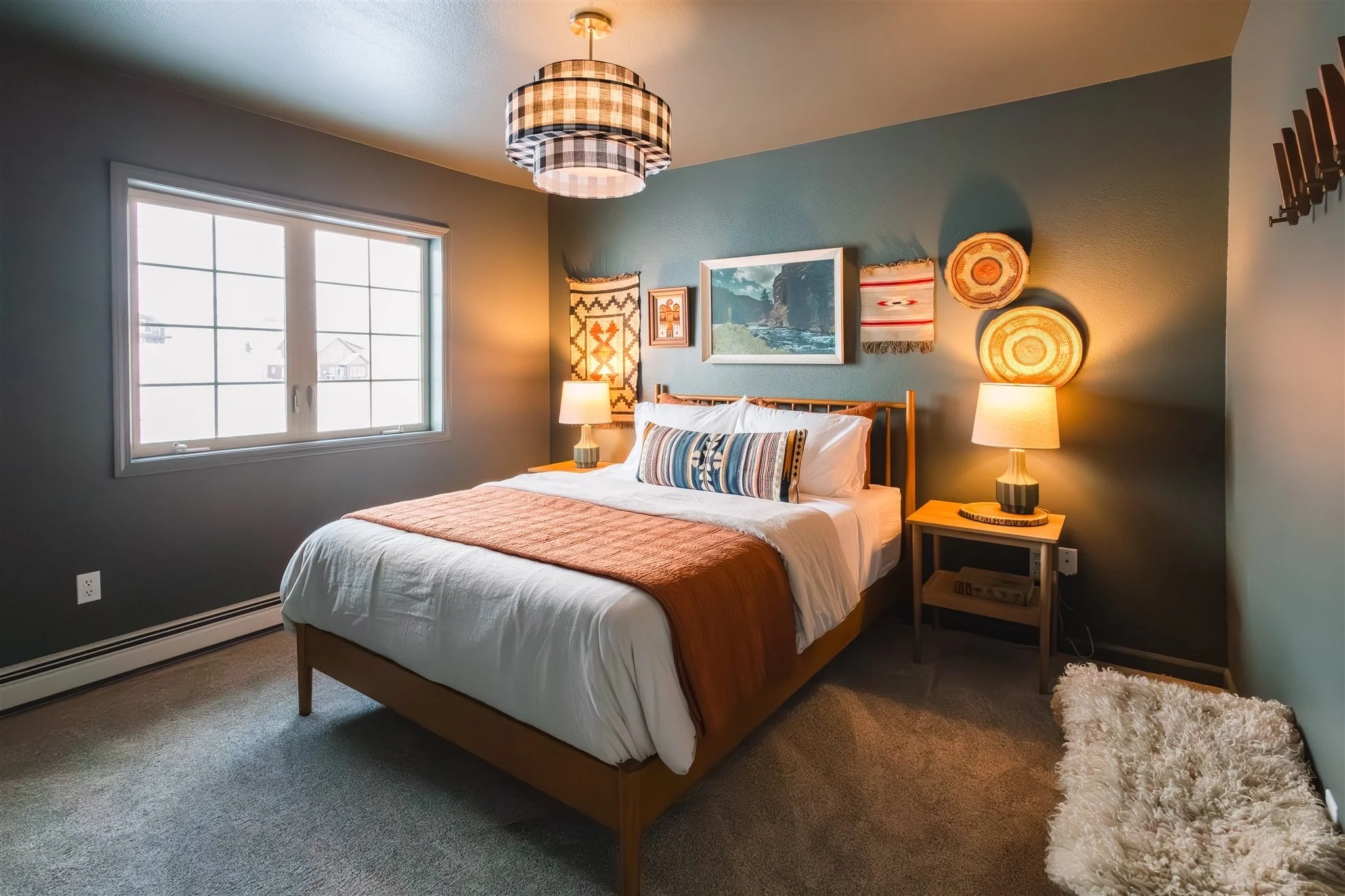 A cozy bedroom with a large window, a bed with a striped pillow and warm bedding, and two nightstands with lamps. The wall has framed artwork and woven decorative pieces. The ceiling has a checkered pendant light.