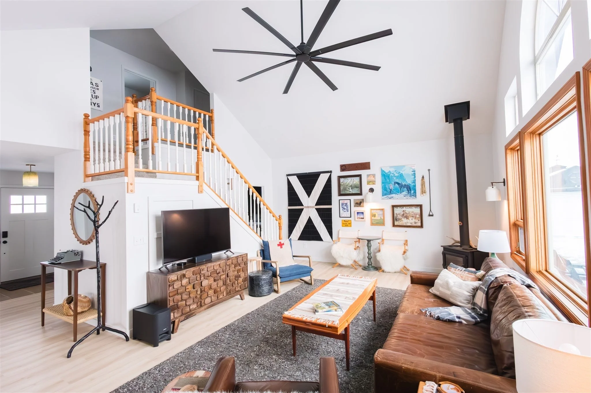 Living room with vaulted ceiling, black modern ceiling fan, large windows, wooden trim, gallery wall with various pictures, wood stove, flat screen TV, leather sofa, chairs with sheepskin throws, coffee table, and various decor items.