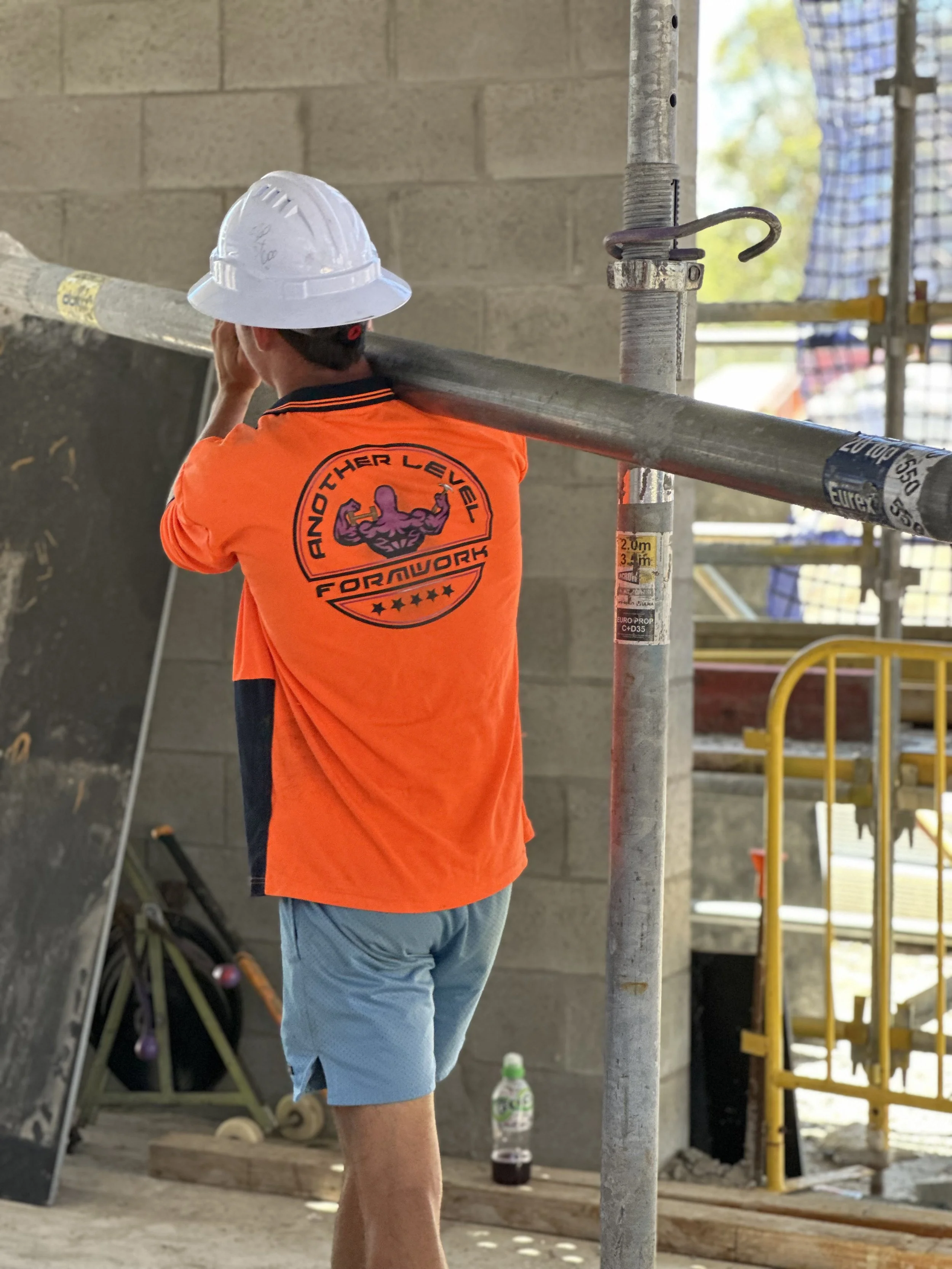 A construction worker wearing an orange shirt with a bodybuilding logo and a white safety helmet, standing at a construction site with scaffolding, a water bottle on the floor, and a gray wall in the background.