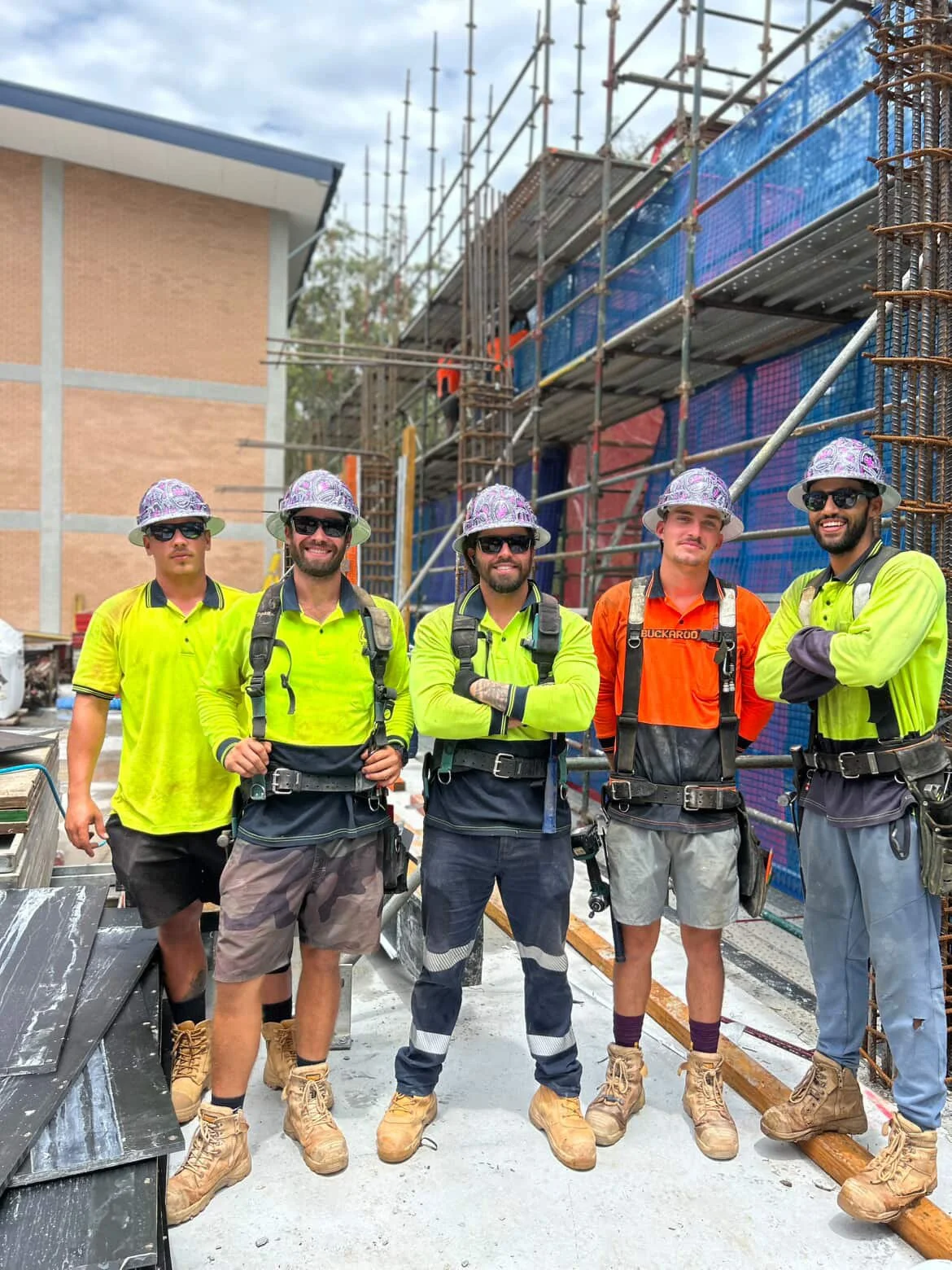 Five construction workers wearing safety gear, including hats, sunglasses, and high-visibility shirts, standing in front of a building under construction with scaffolding.