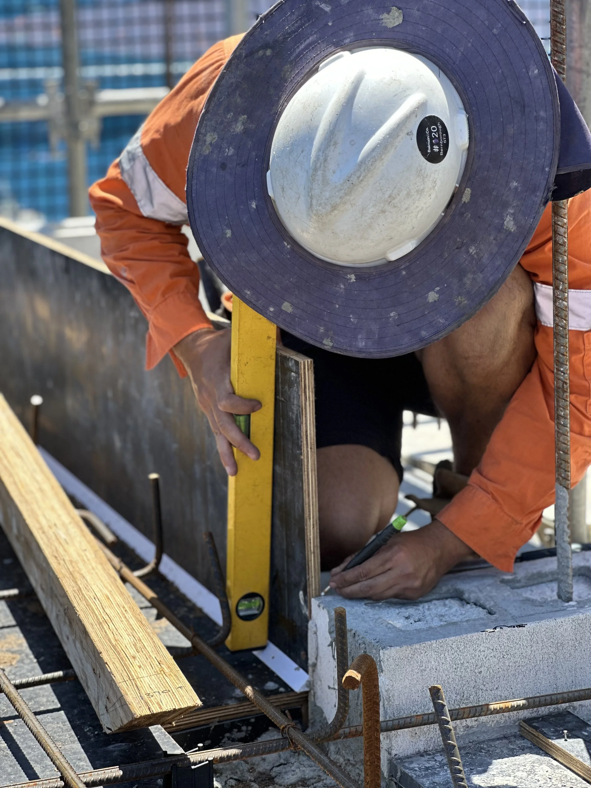 A construction worker wearing an orange shirt and a white safety helmet, holding a level and working on a concrete structure at a construction site.