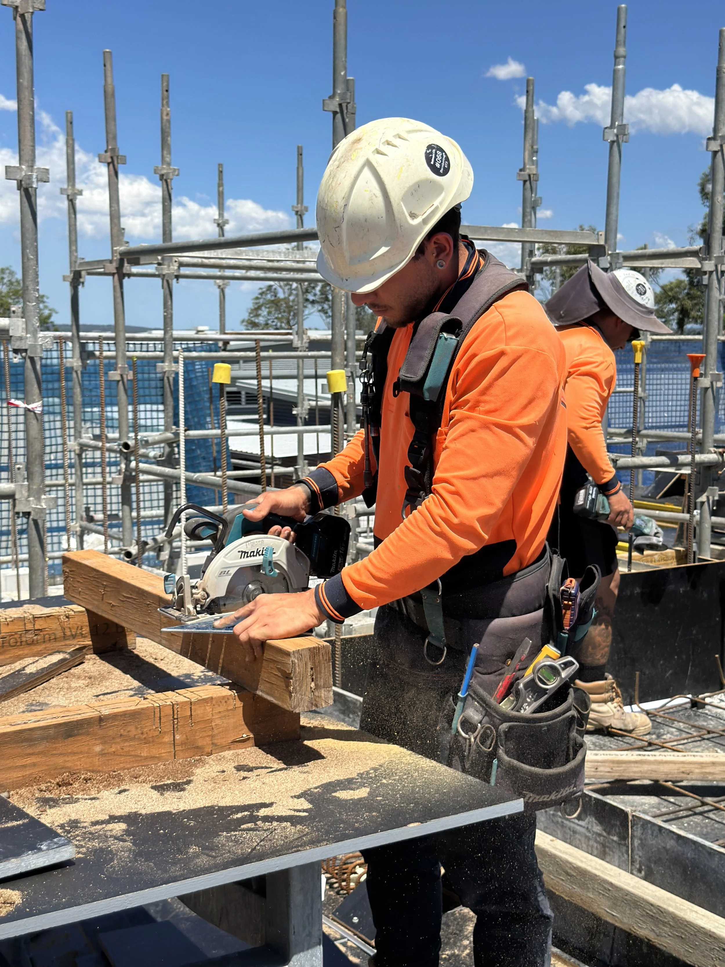 A construction worker wearing an orange safety jacket, a white helmet, and a tool belt, is operating a chainsaw to cut a wooden beam at a construction site with scaffolding and safety barriers under a blue sky.