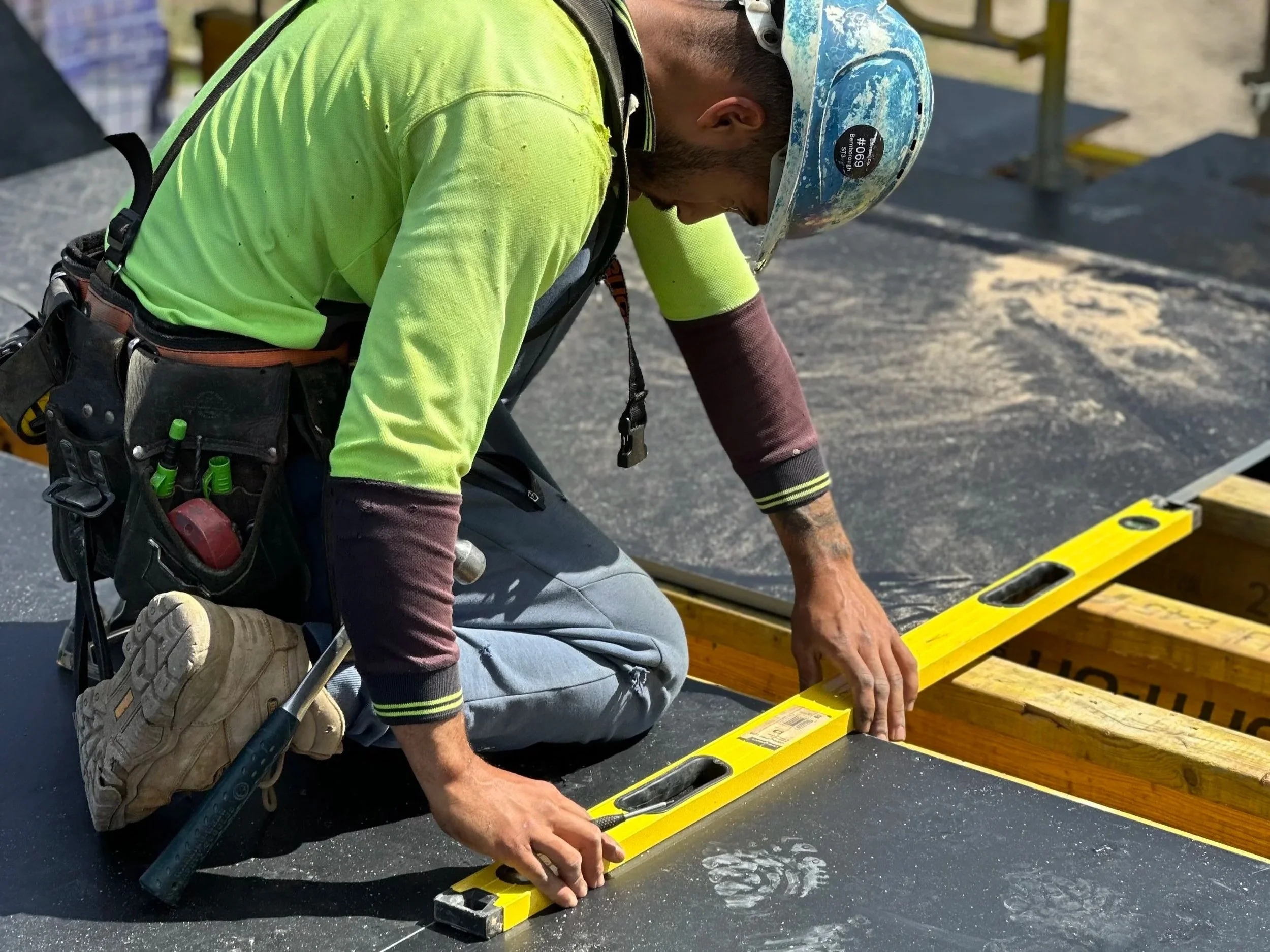 A man wearing a bright yellow safety shirt, a blue helmet with safety stickers, and work gear kneels on the ground, measuring and working on a yellow wooden frame or platform outdoors.