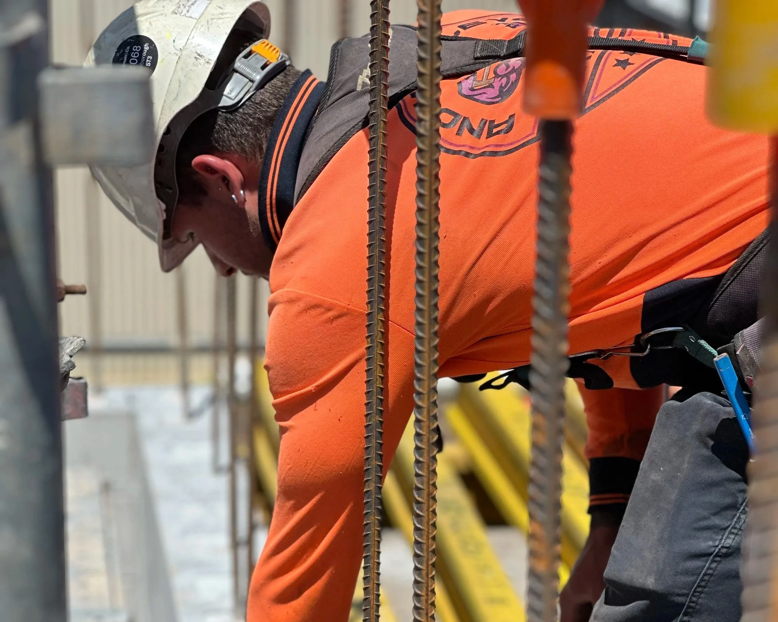 Worker in orange safety clothing leans forward while working with electrical or construction materials behind a safety barrier.