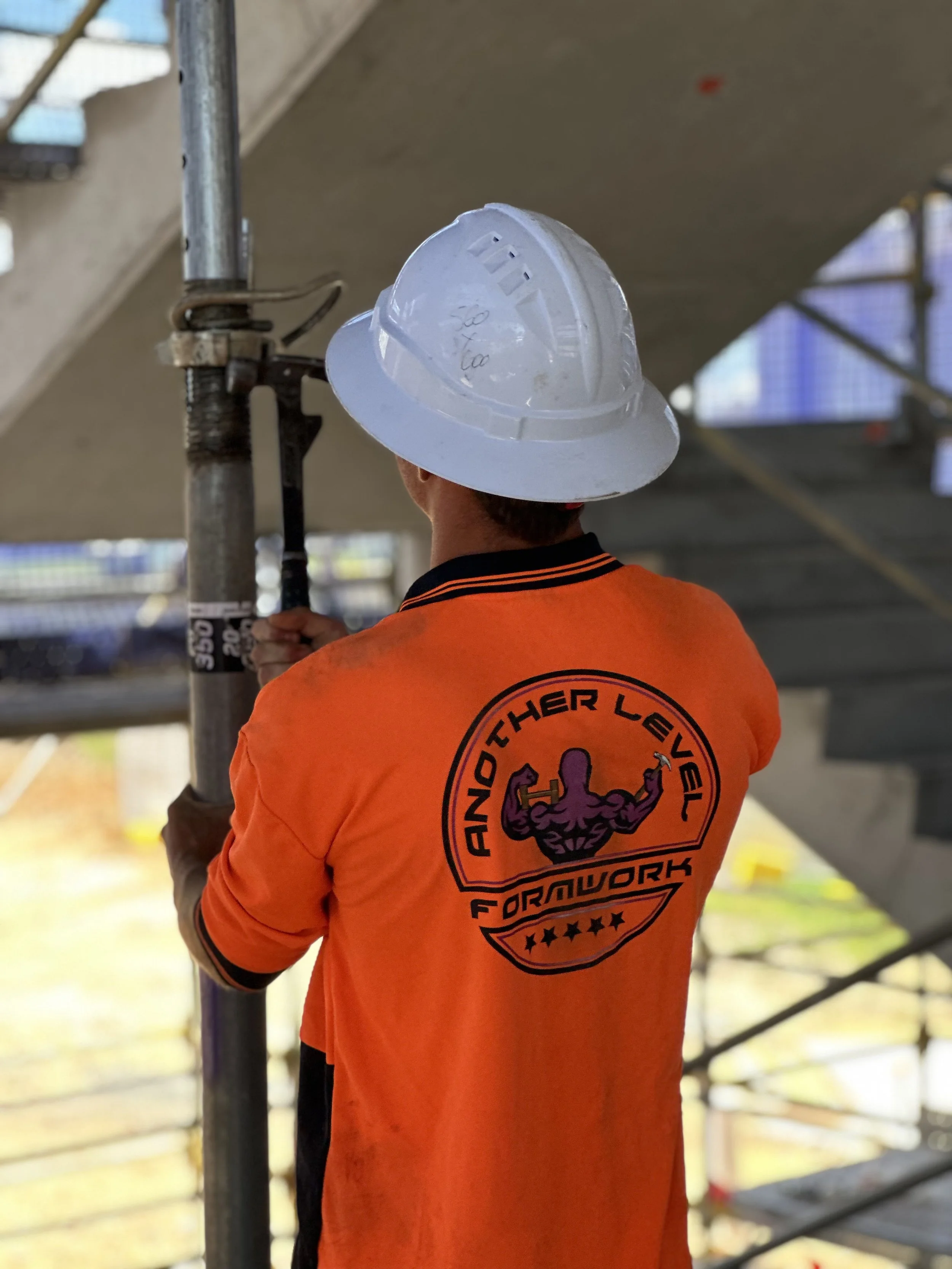 A construction worker wearing an orange shirt with a logo on the back, and a white hard hat, is standing on a construction site near scaffolding.