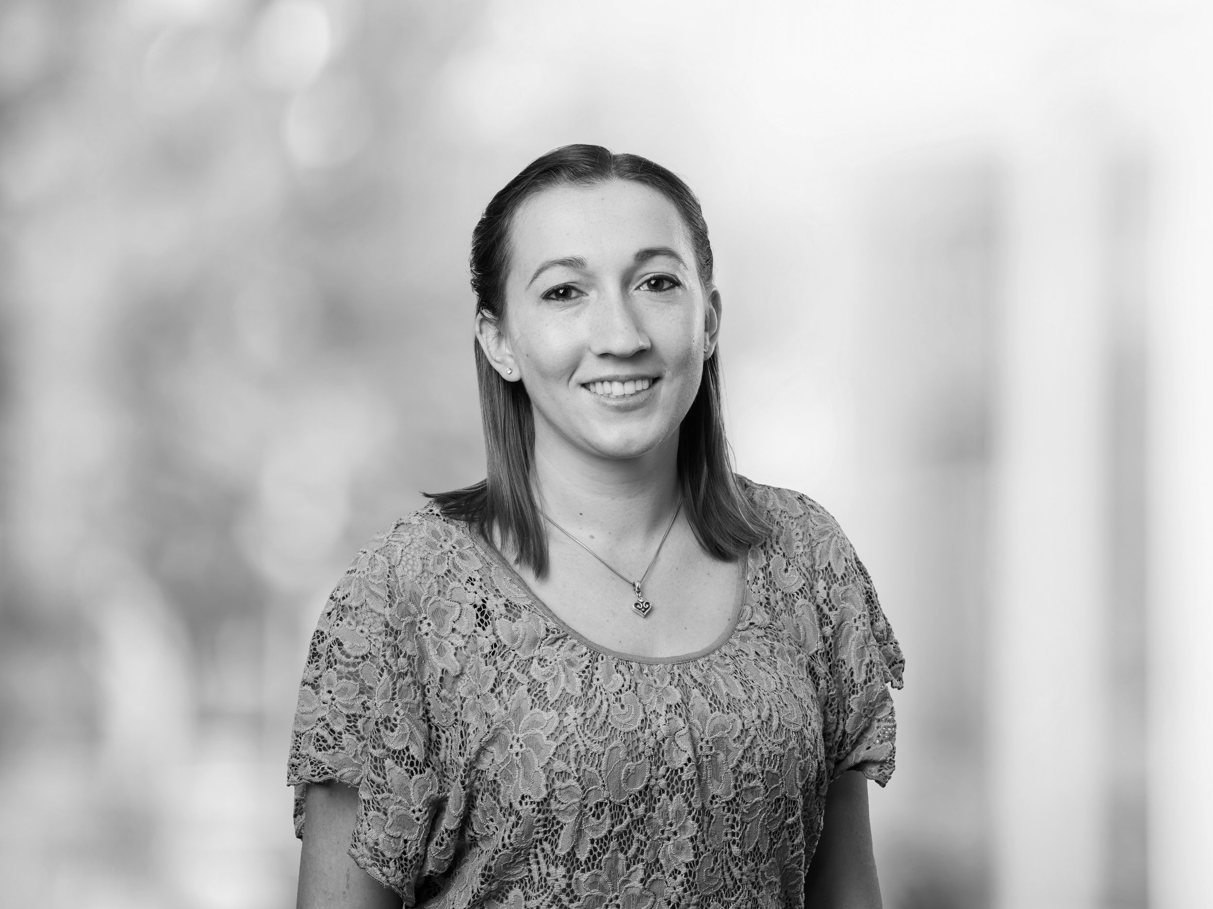 Black and white portrait of a smiling woman with straight hair wearing a lace top and a necklace with a heart-shaped pendant, standing against a blurred background.