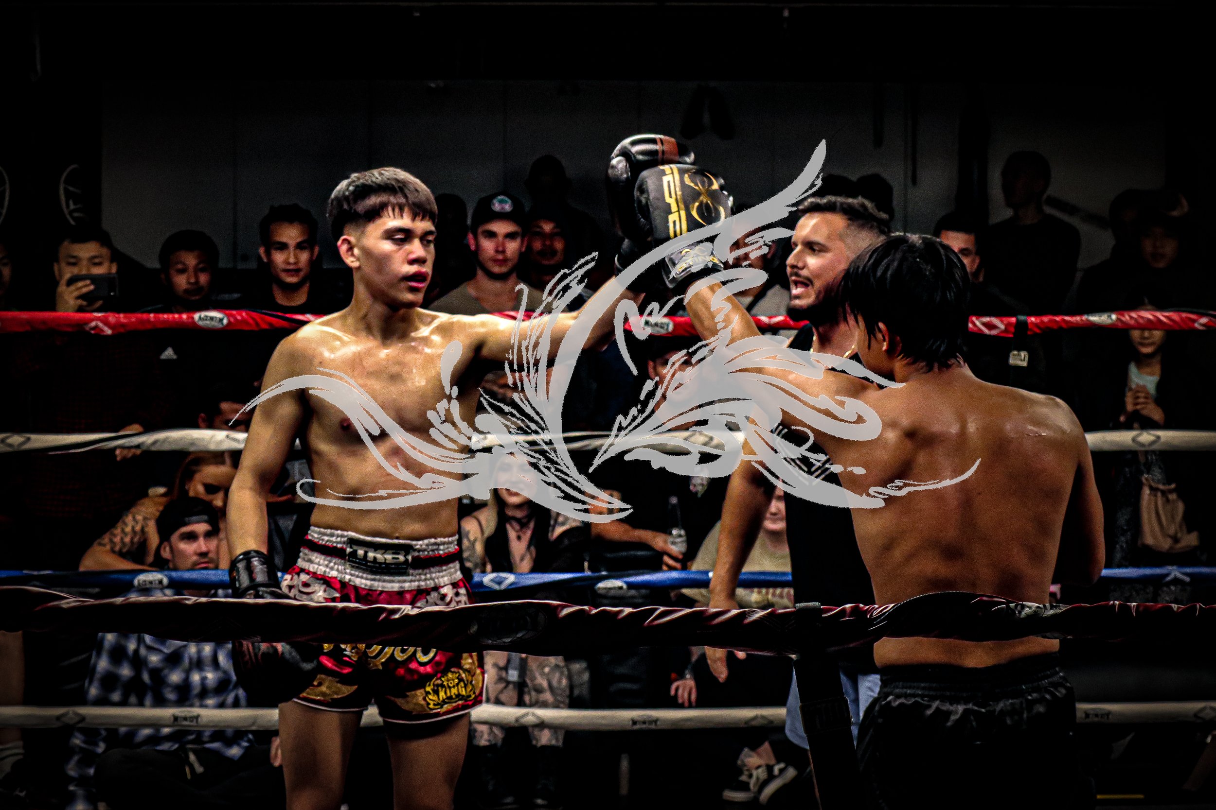 Two shirtless Muay Thai fighters in a ring, one with a black and red helmet and gloves, the other with black gloves, surrounded by spectators watching the match.