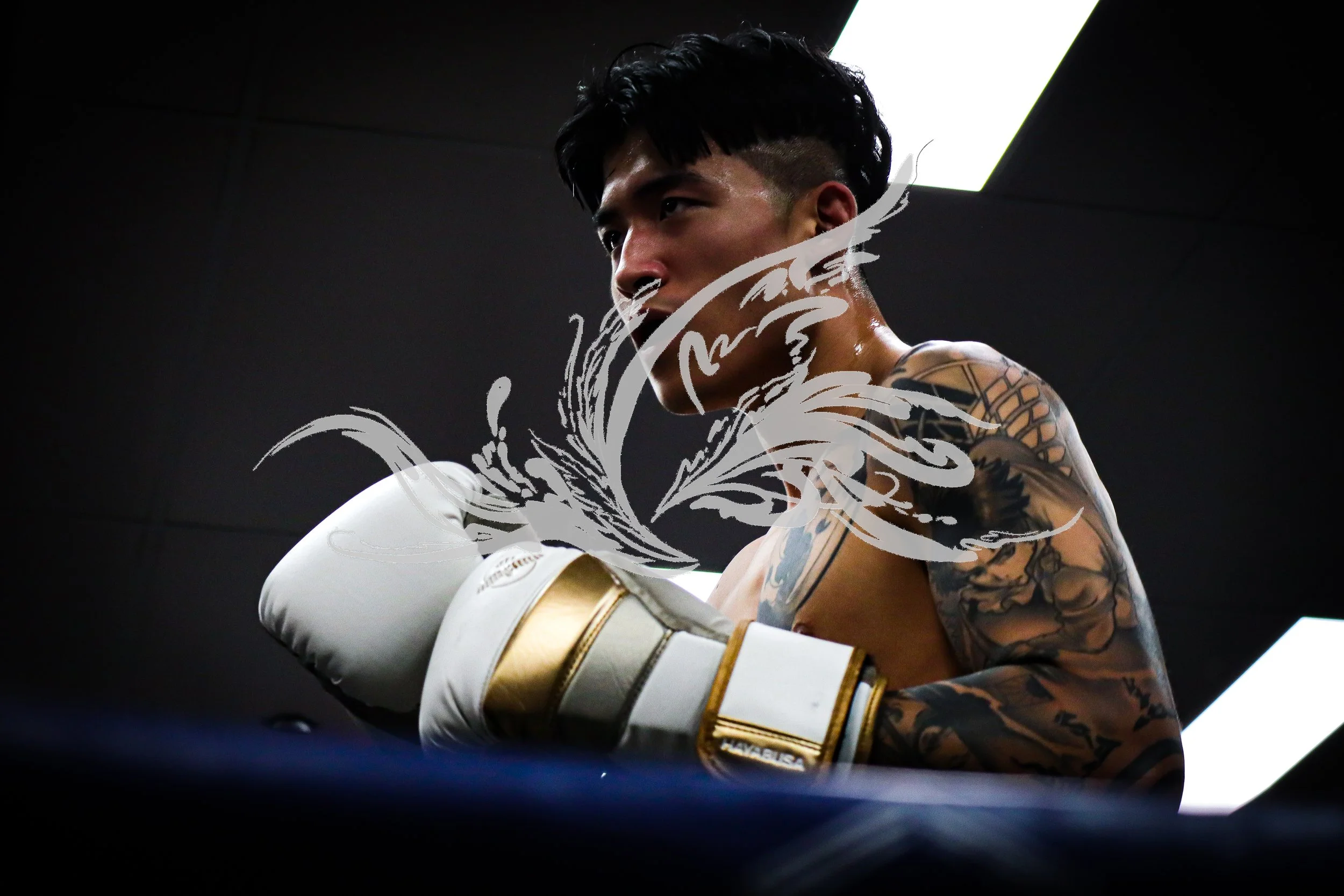 A shirtless male boxer with tattoos wearing white and gold boxing gloves, standing in a boxing ring with low lighting and a dark background.
