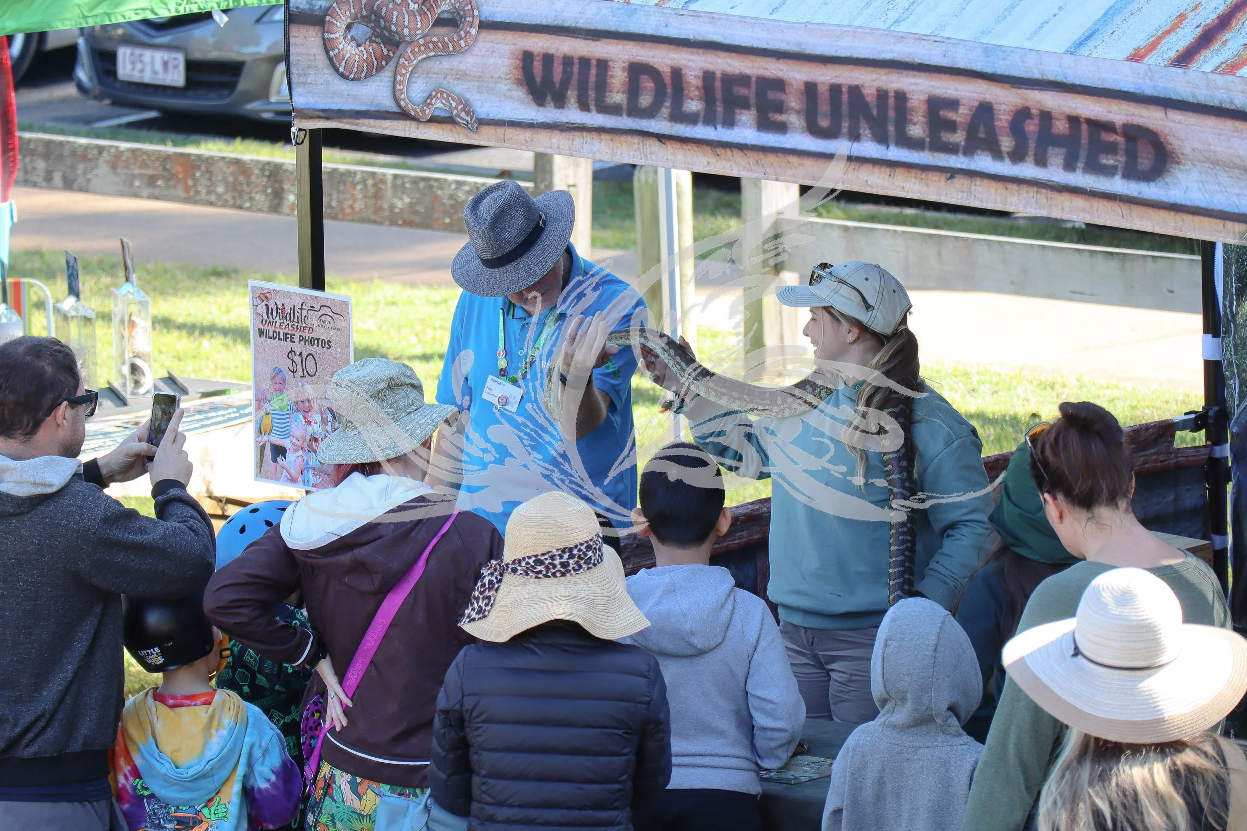 People gathered around a booth with a banner that says 'Wildlife Unleashed'. The booth features a snake presentation, with a person holding a snake. The crowd includes children and adults, some taking pictures. The setting appears to be outdoors, pos