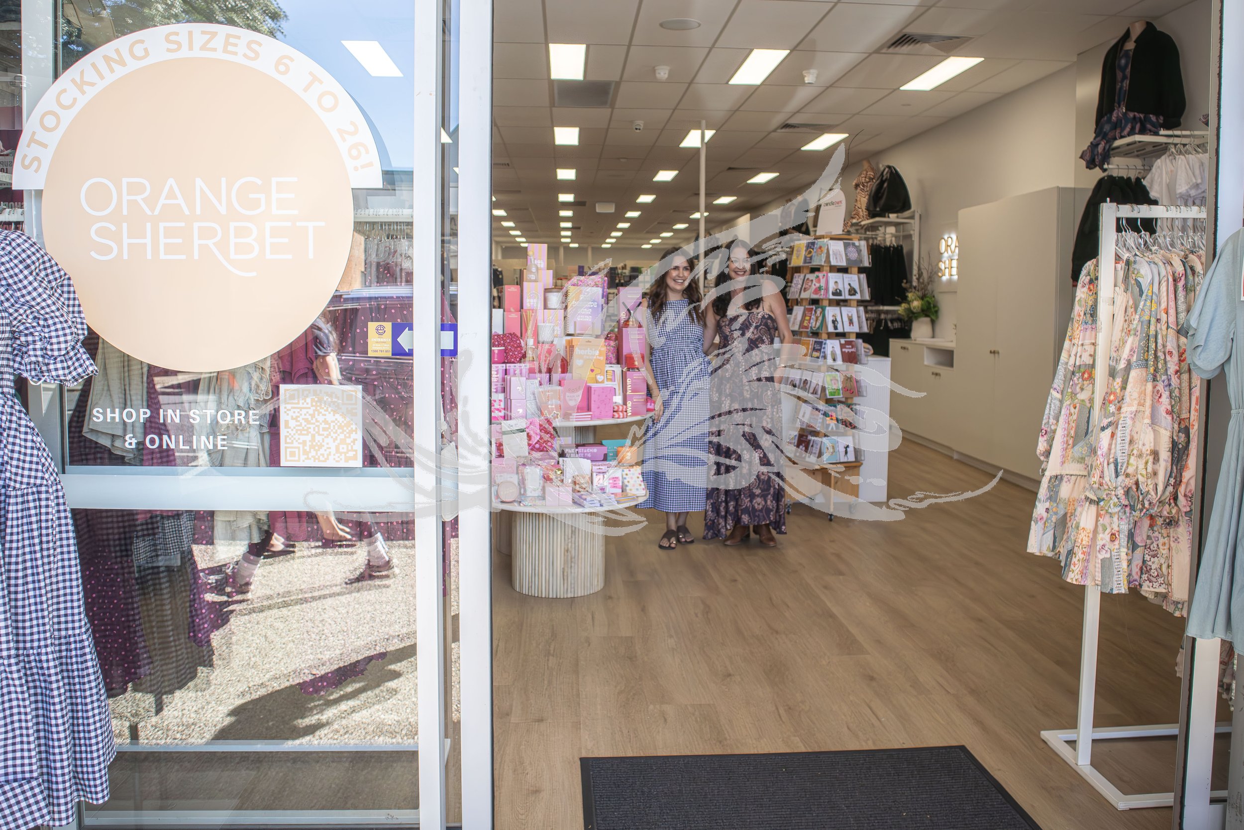 View inside a retail store with two women smiling at the entrance, surrounded by clothing and accessories displayed on racks and shelves, with bright lighting and a wooden floor.