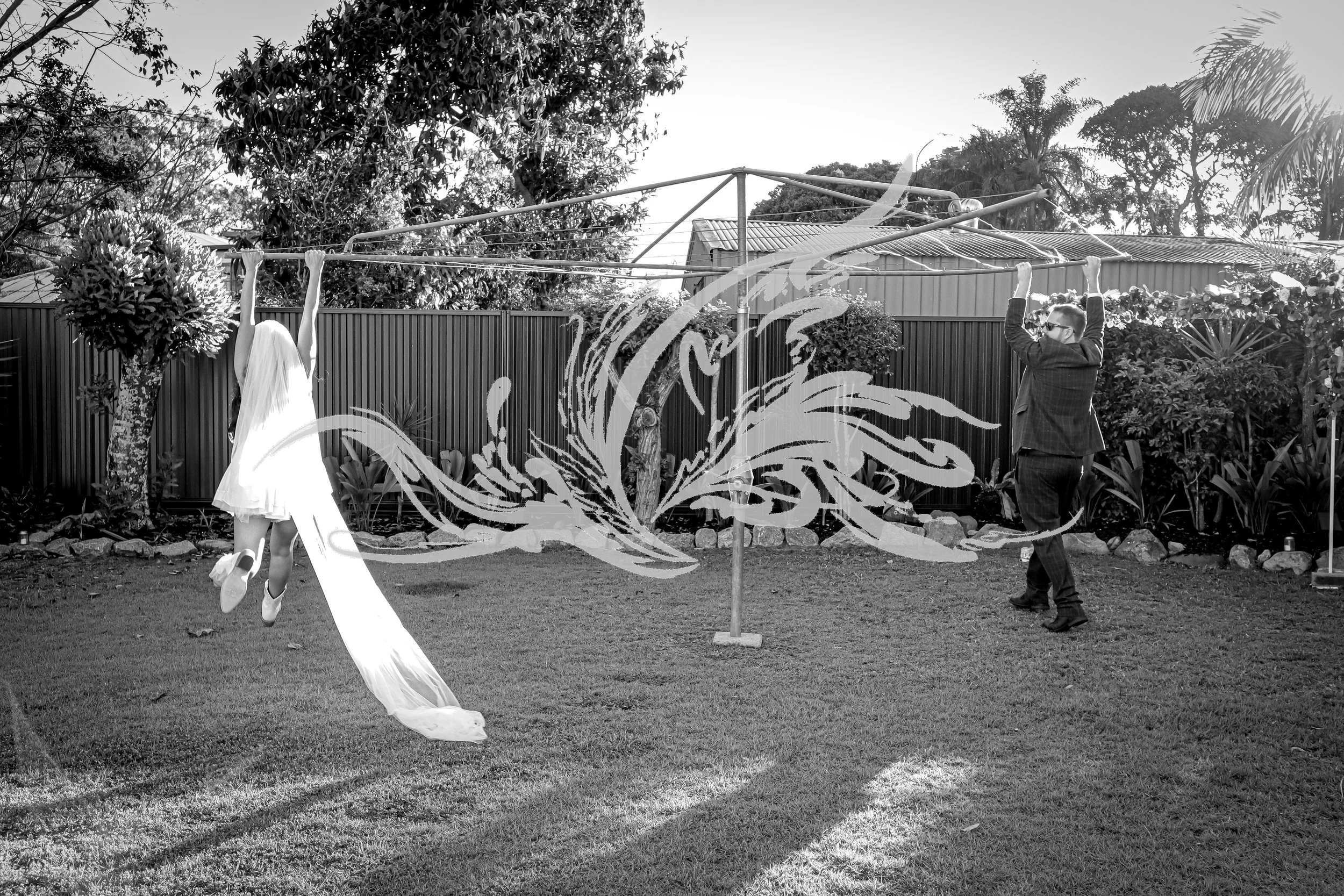 A black and white photo of a woman in a white dress jumping in the air while a man in a suit throws a bouquet into the air in a backyard with a clothesline, trees, and a shed in the background.