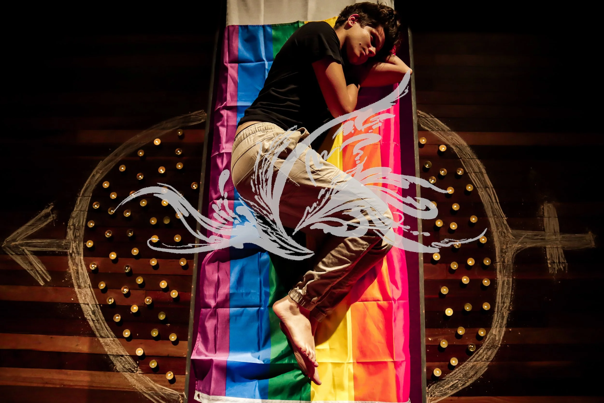 A person lying on a rainbow flag on a stage, with a rainbow Pride flag draped over a rainbow-colored podium, surrounded by a decorative heart-shaped outline with small lights.