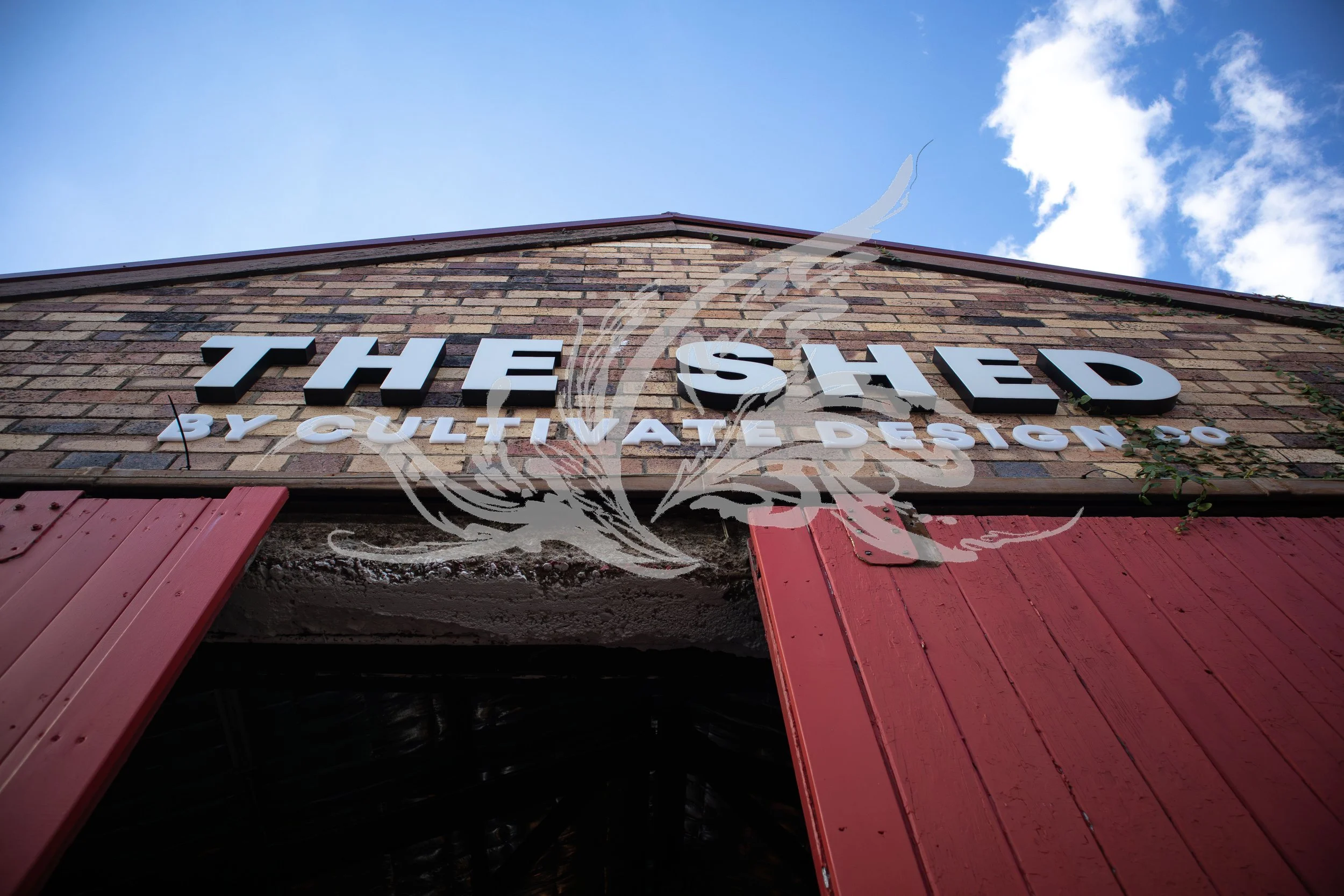 View of the exterior brick wall of a restaurant called "The Shed" with red wooden accents and a cloudy blue sky in the background.