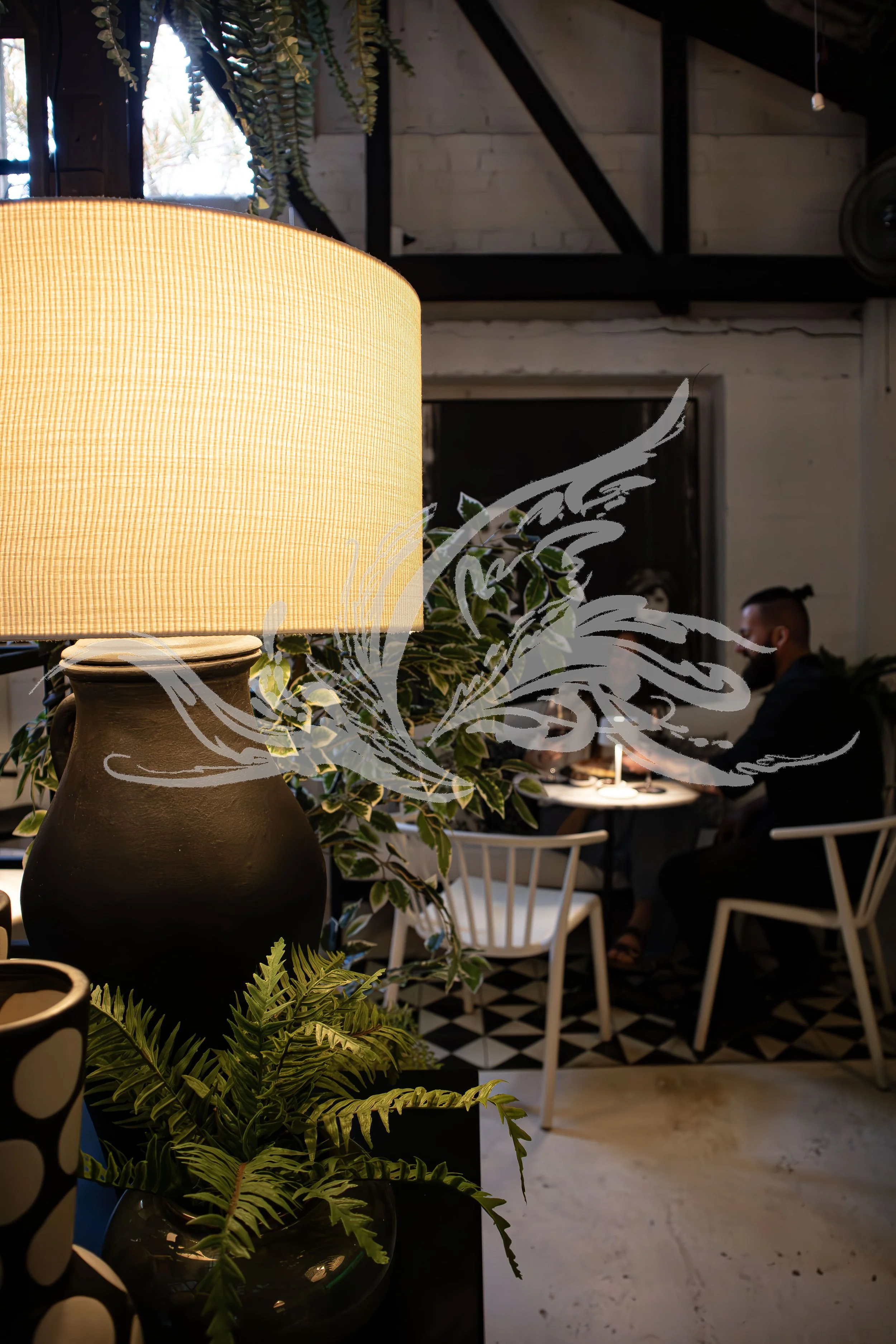 Interior of a stylish cafe or restaurant with warm lighting, plants, and white chairs, with a person in the background.
