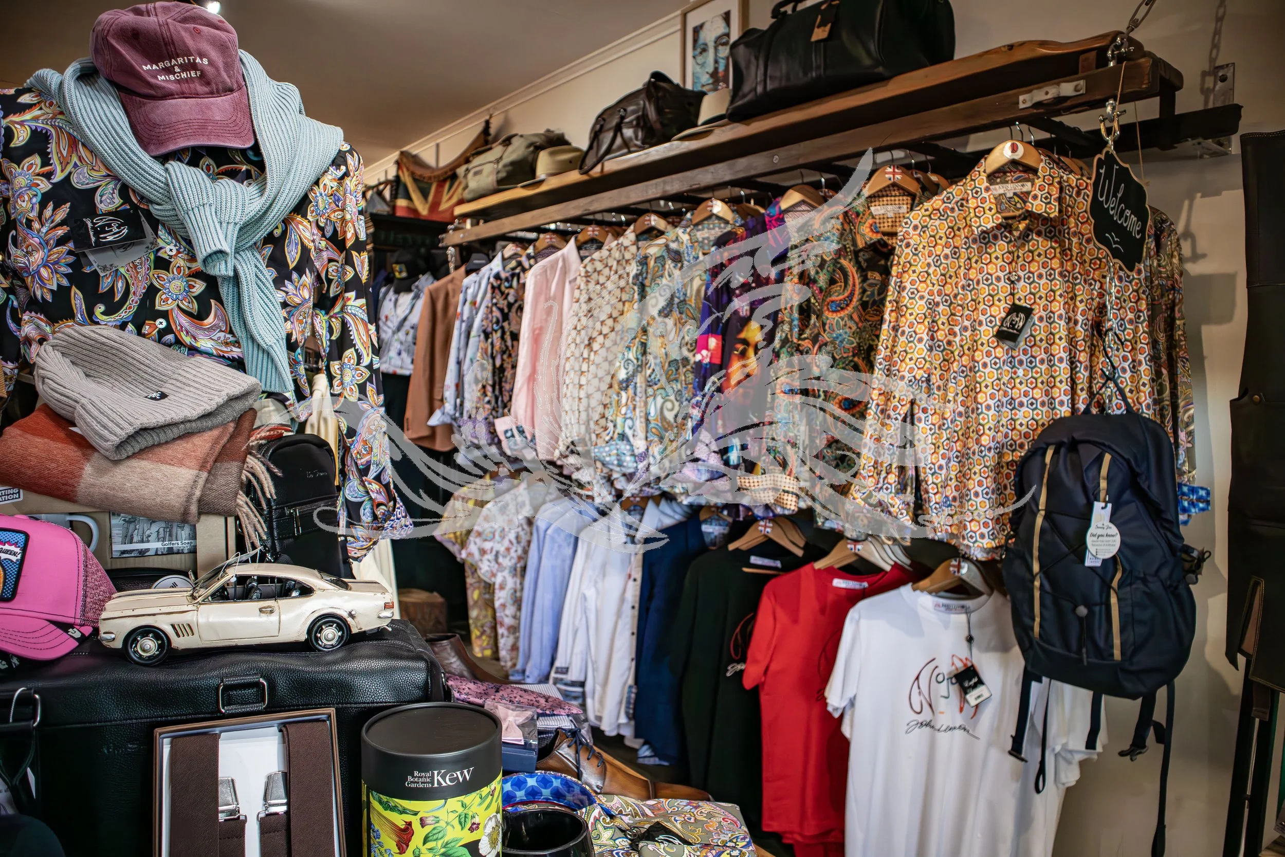 A variety of secondhand clothing, accessories, and household items are displayed on shelves and tables at a thrift store. Includes patterned shirts, hats, bags, a toy car, and miscellaneous decor items.