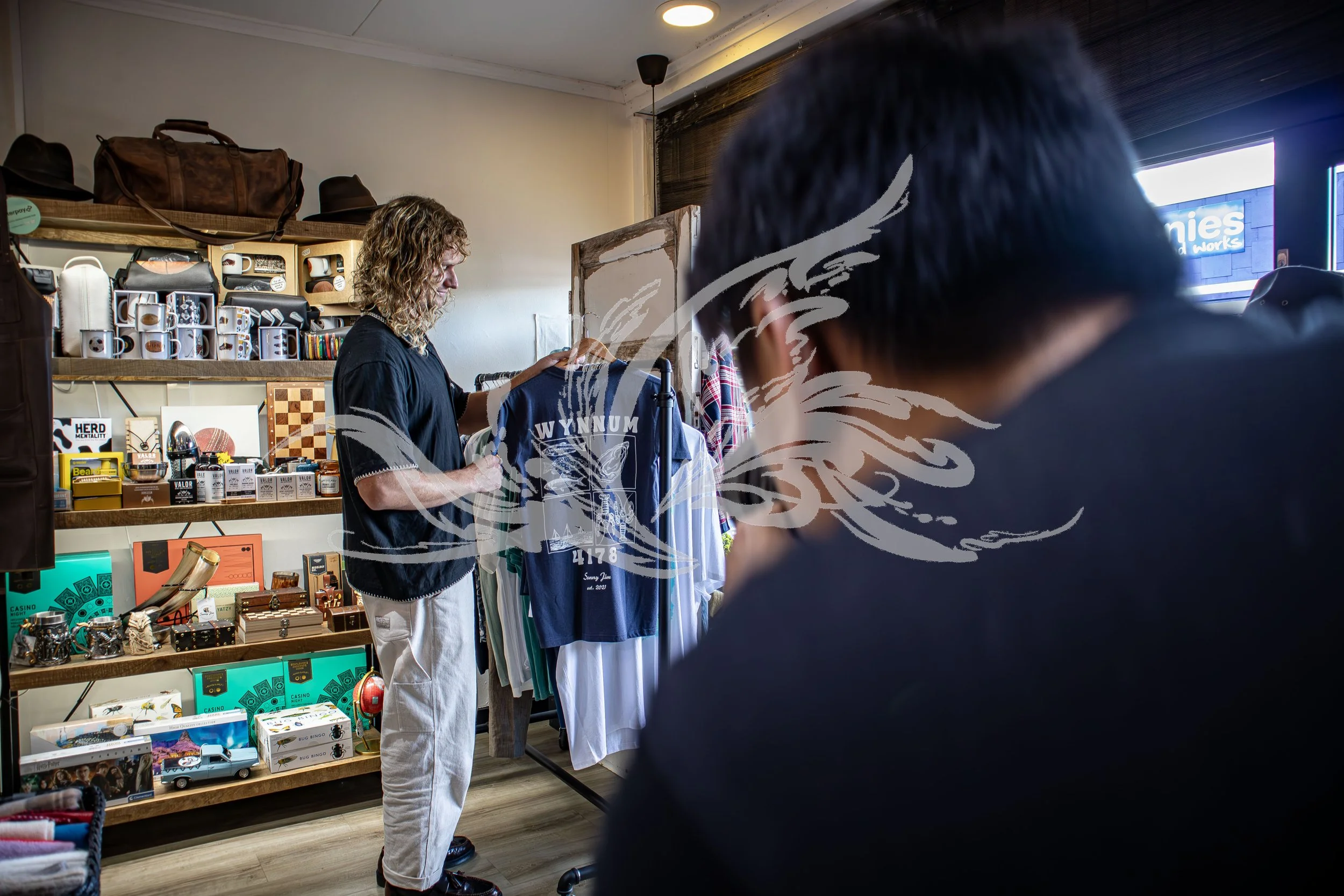 Inside a store with shelves of mugs, souvenirs, and accessories. A woman with curly hair looks at clothes on a rack. The store has a wooden floor and a white wall, with hats and bags on shelves. Part of a person in dark clothing is visible in the for