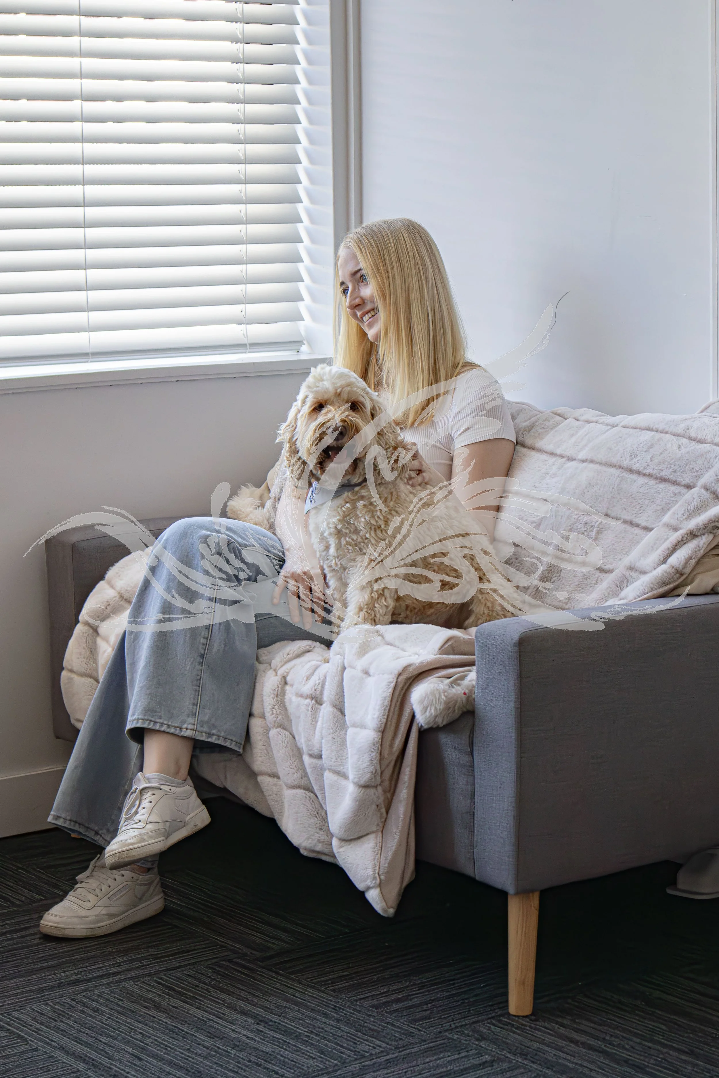 A young woman with blonde hair sitting on a bed in front of a window with white blinds, holding a large fluffy dog. She is smiling and wearing beige sneakers, a light-colored striped shirt, and loose gray jeans. The bed has a plush blanket and is nex