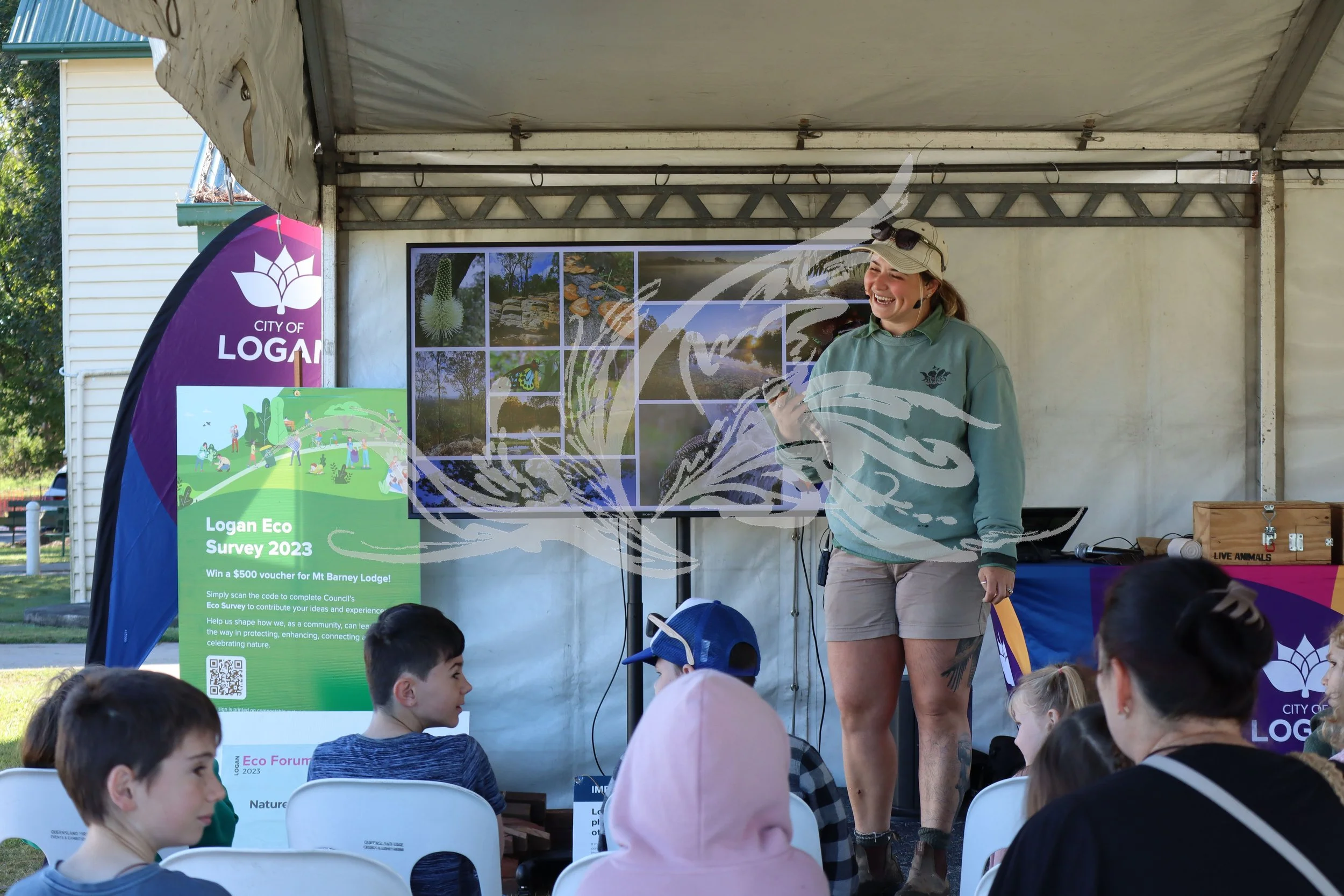 A group of children attending the Logan Eco Forum 2023, seated and listening to a woman speaker under a tent. The woman is smiling and holding a microphone, with eco-related posters and signs around her.