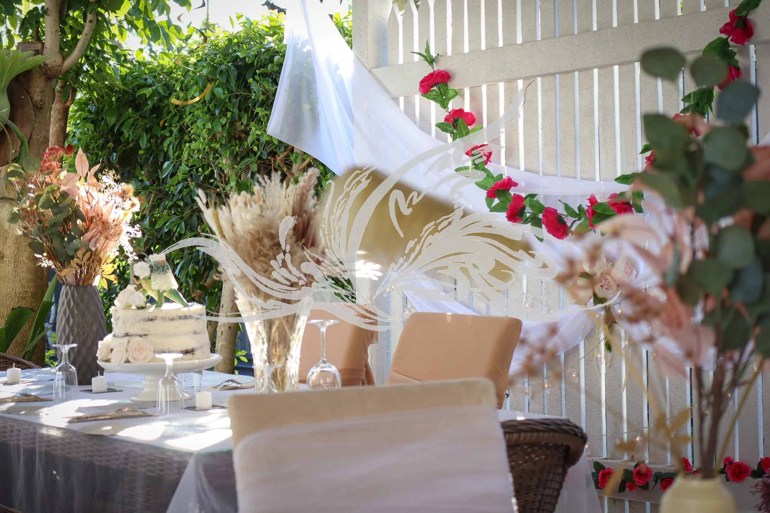 Outdoor event decorated with pink flowers and greenery, includes a table with a cake, glassware, and floral arrangements, under a white draped fabric on a patio or garden setting.