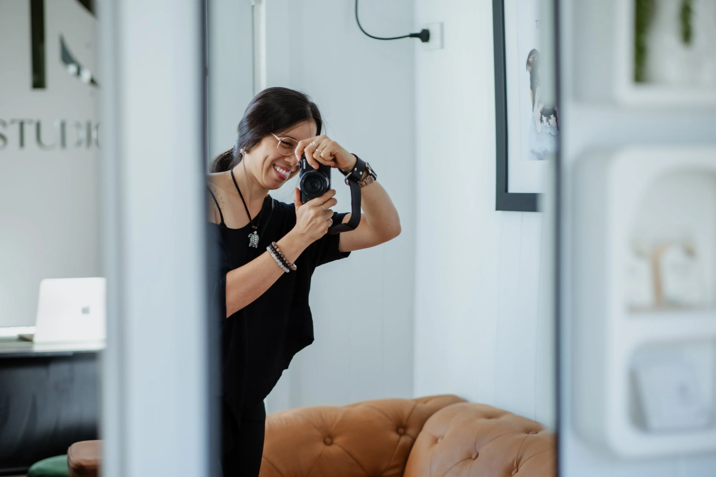 A woman smiling and taking a photo with a camera in a bright room, reflected in a mirror, with a tan leather sofa in the background.