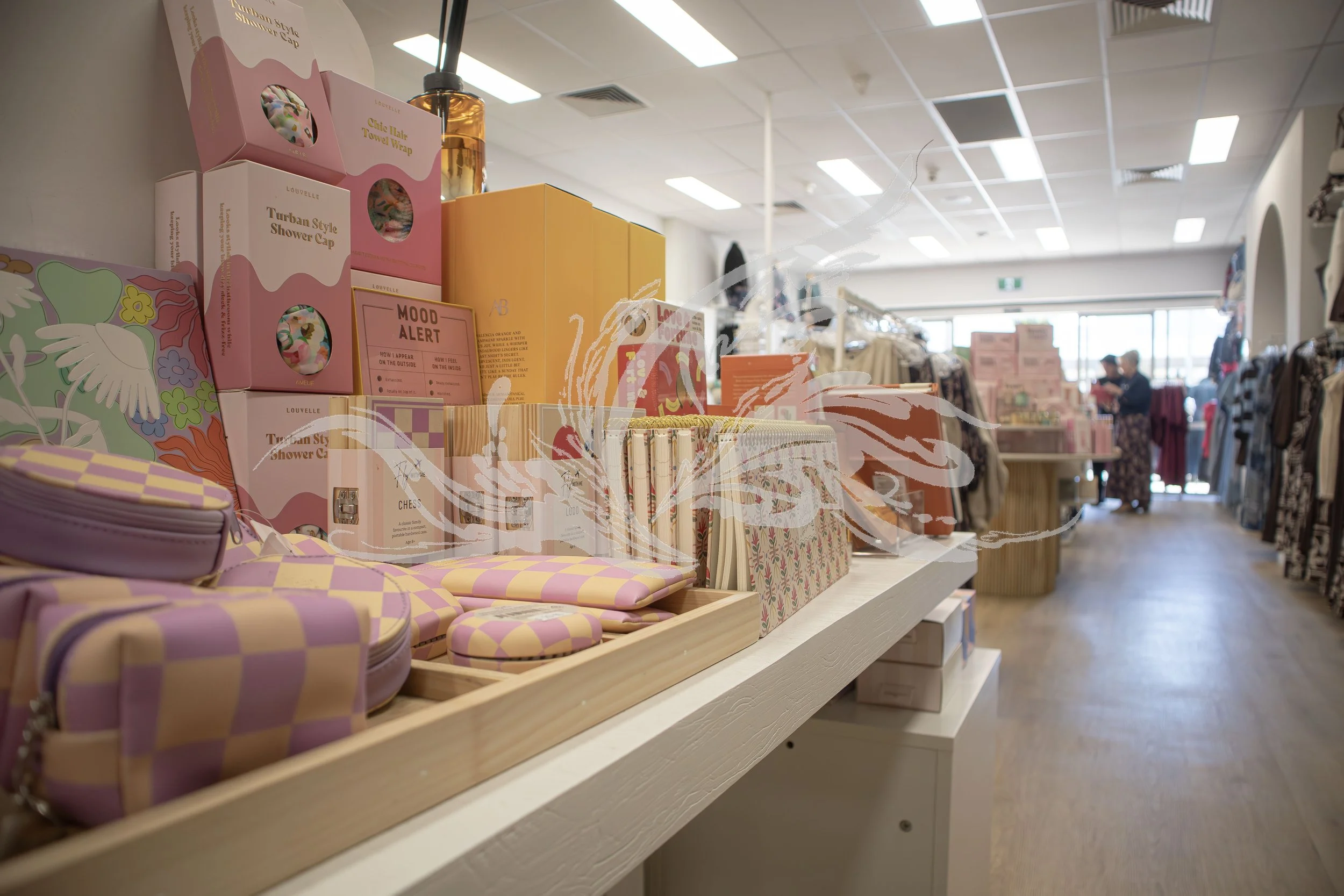 A store shelf displays colorful slippers with a pink and yellow checkered pattern, along with other miscellaneous items like notebooks and packaging, in a retail store with clothing racks and customers in the background.