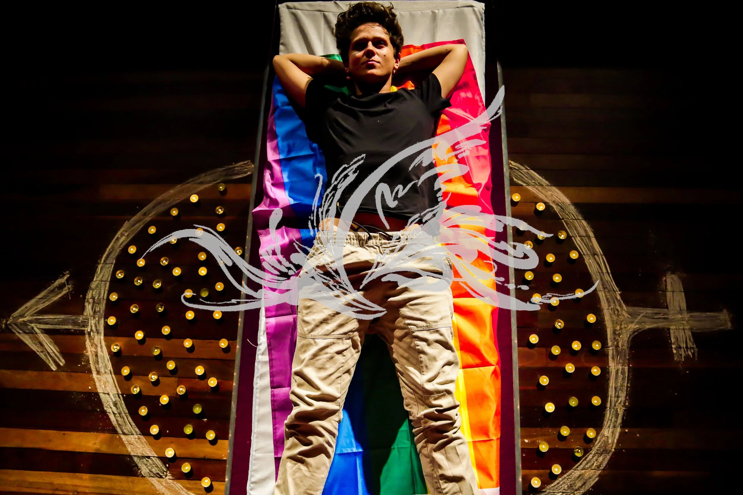 A young man lying on a colorful rainbow pride flag on a star-shaped bed with a wooden backboard decorated with yellow lights, looking up with arms behind his head.