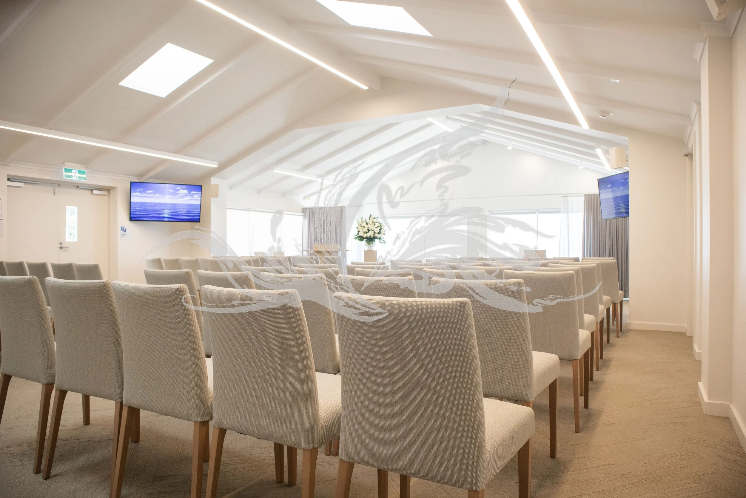 Empty conference room with rows of beige chairs, two wall-mounted screens displaying a blue sky and ocean scene, potted plant, curtains, and a white ceiling with modern lighting.