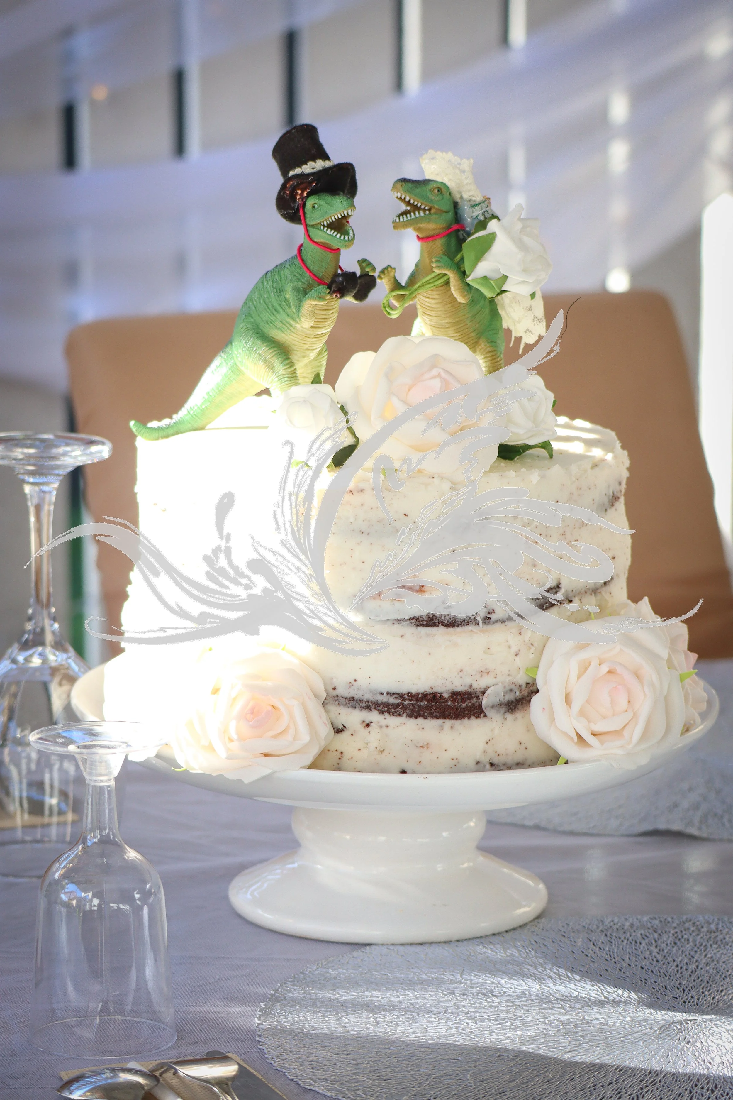 A wedding cake decorated with white flowers and two dinosaur figurines dressed as a bride and groom, placed atop a white cake stand on a table with glassware and tablecloths.