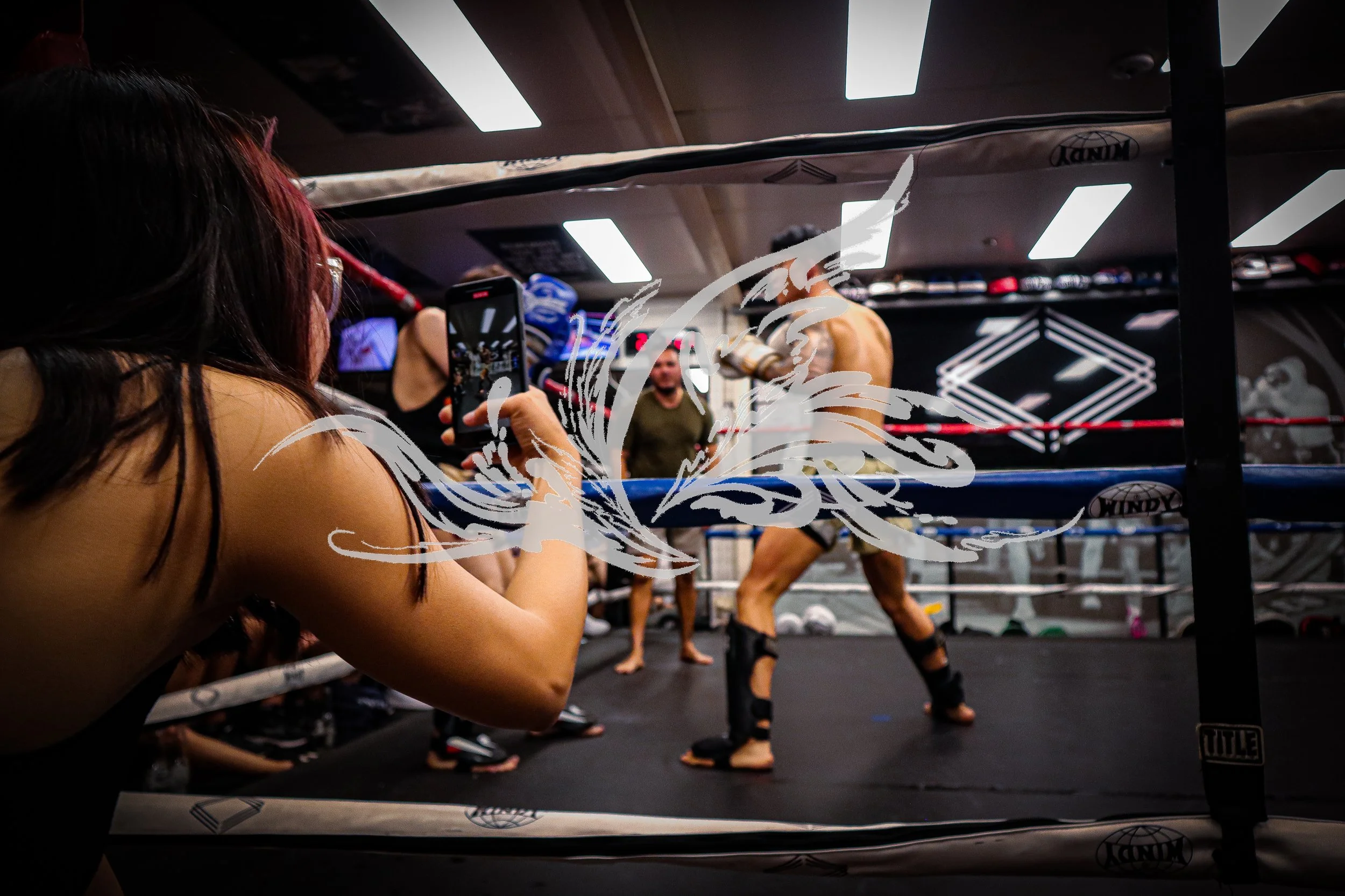 A woman with dark hair and glasses taking a photo or video of a boxing match inside a gym. The boxer is wearing blue gloves and shorts, with another fighter in the background. The gym has bright ceiling lights and boxing equipment.