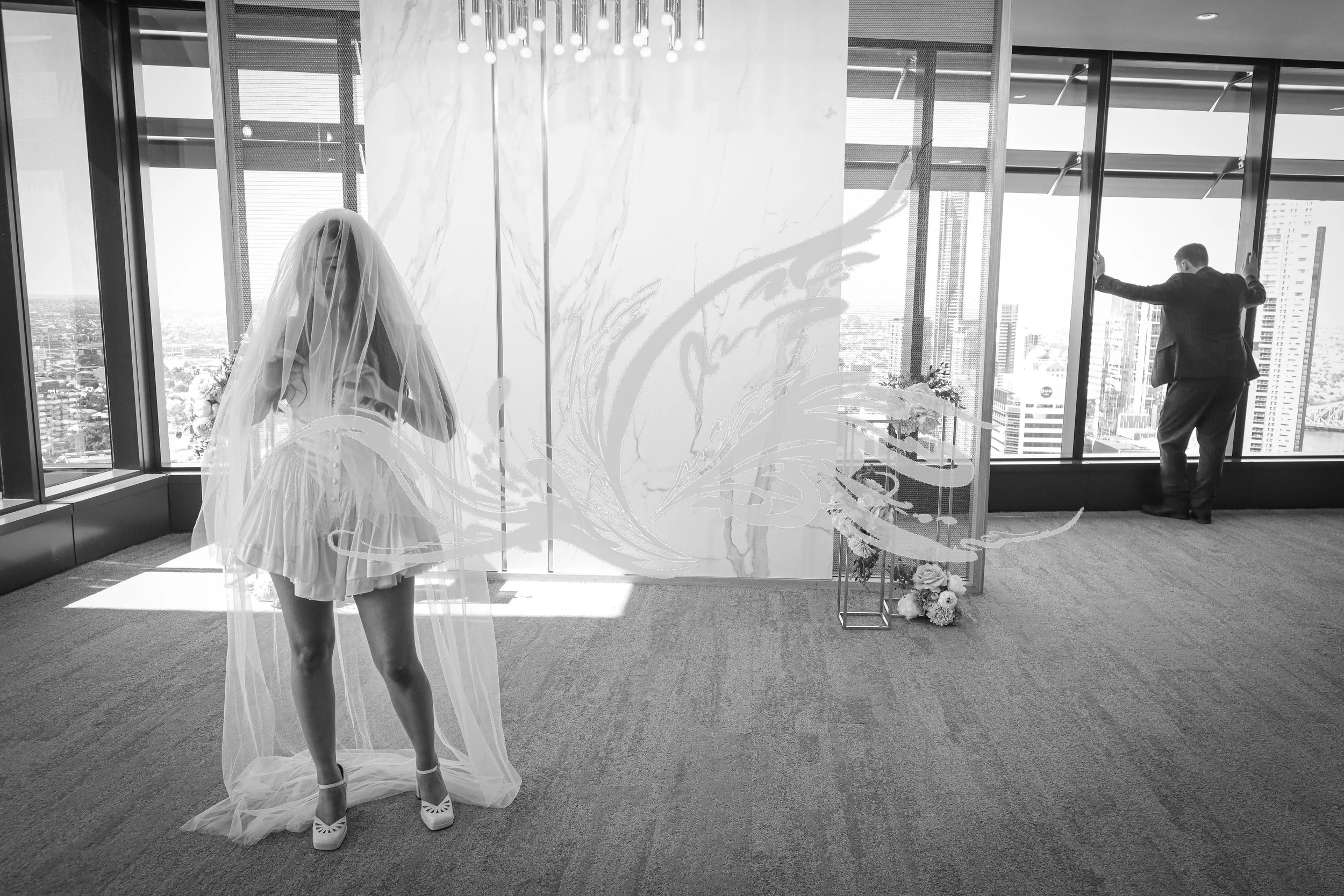 A bride in a wedding dress with a veil stands on a balcony with city skyline views in the background. A man in a suit is adjusting a glass door window. There is a floral arrangement and a decorative wall with lighting fixtures inside the room.