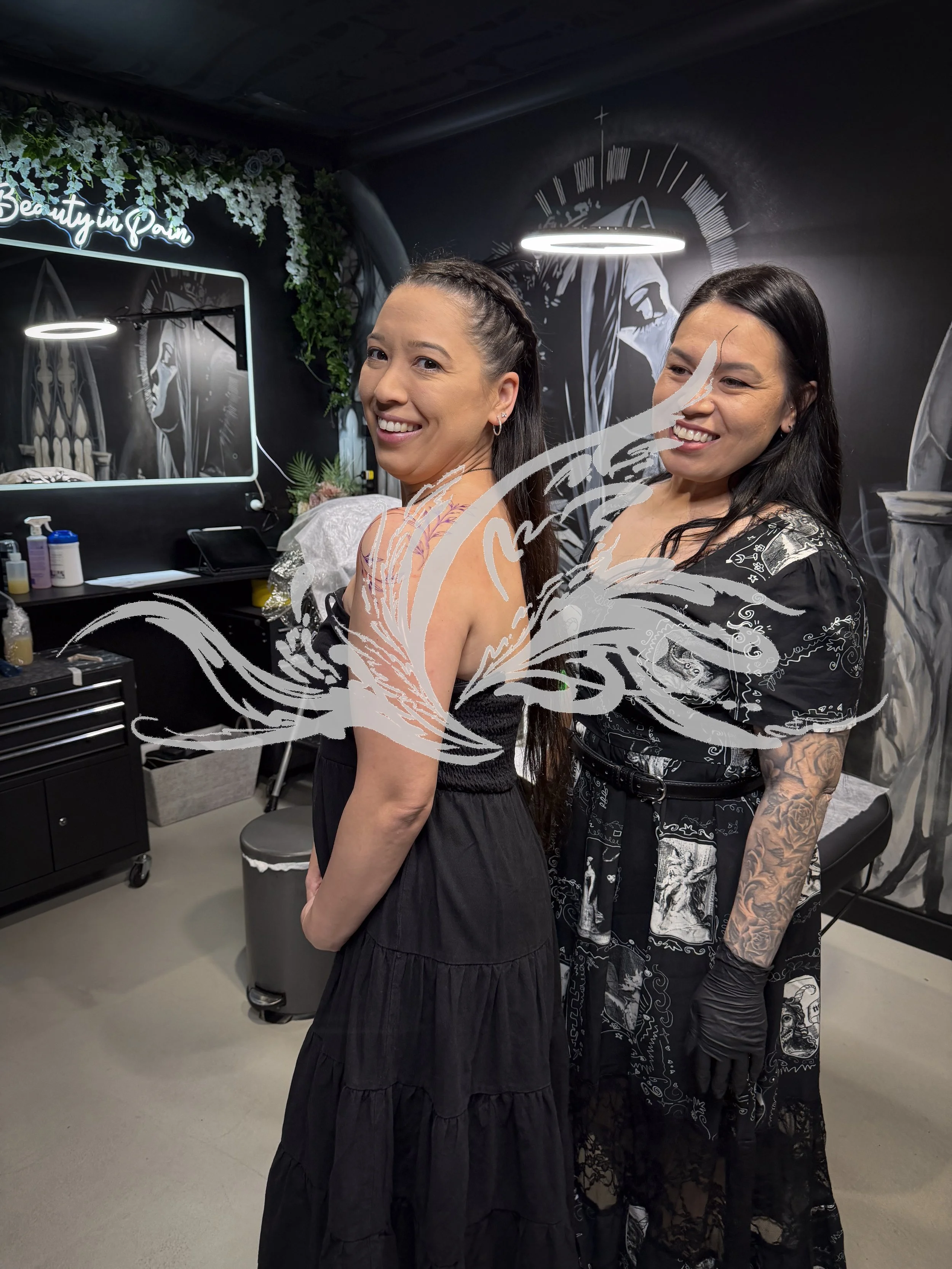 Two women in gothic-inspired attire standing in a tattoo studio with dark walls, tattoo art, and soft lighting, smiling at the camera.