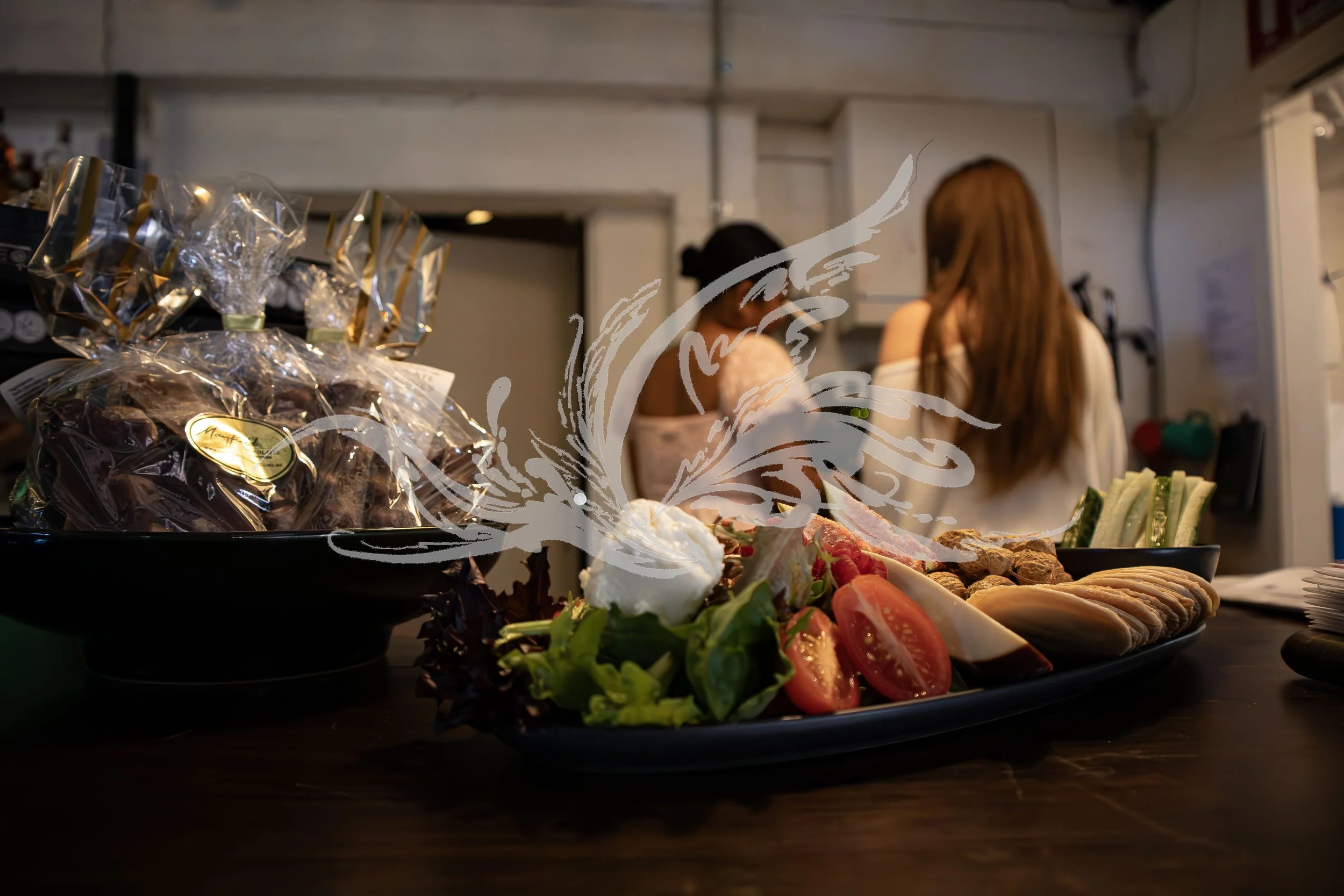 A table with a platter of sliced vegetables and meats, and a smaller bowl of wrapped candies in the background, with two women chatting in a kitchen setting.