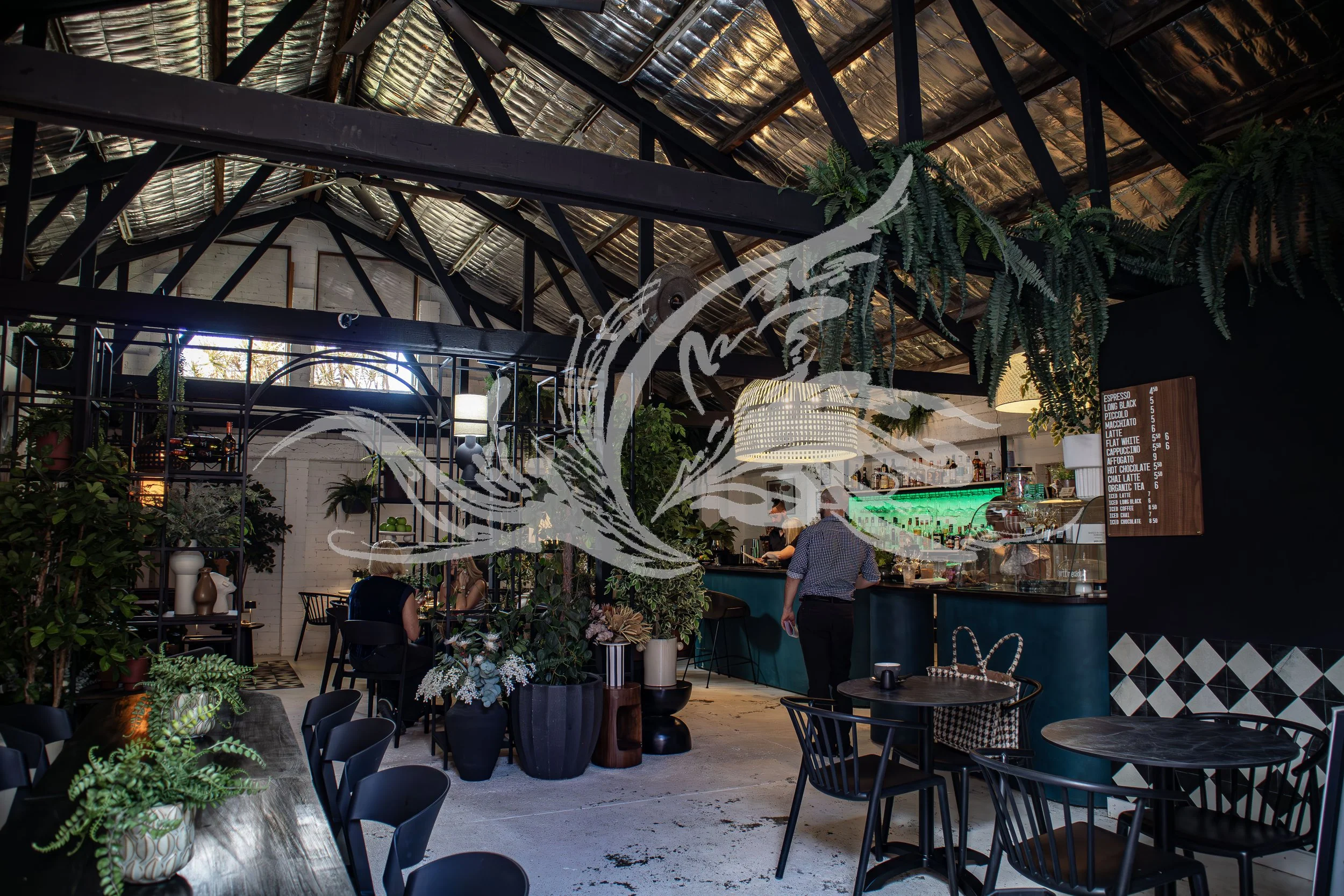 Interior of a modern cafe with black chairs and tables, green plants, and a bar counter with a menu board. The ceiling has a rustic, corrugated metal roof with exposed beams and hanging plants.