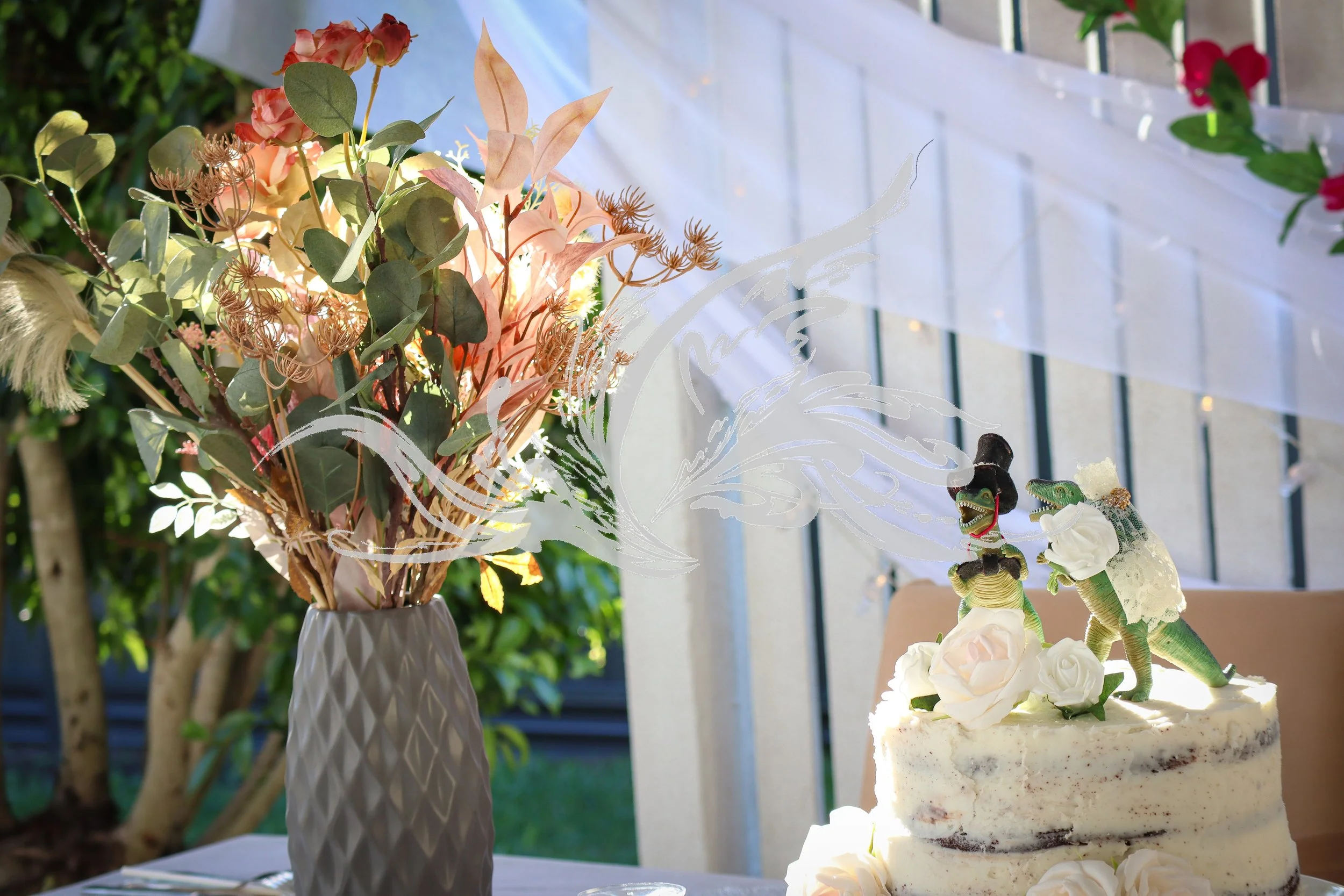 A floral bouquet in a white vase and a two-tiered white cake decorated with two dinosaur figurines and white roses, set on a table with a white tablecloth, outdoors with green foliage and a white railing in the background.