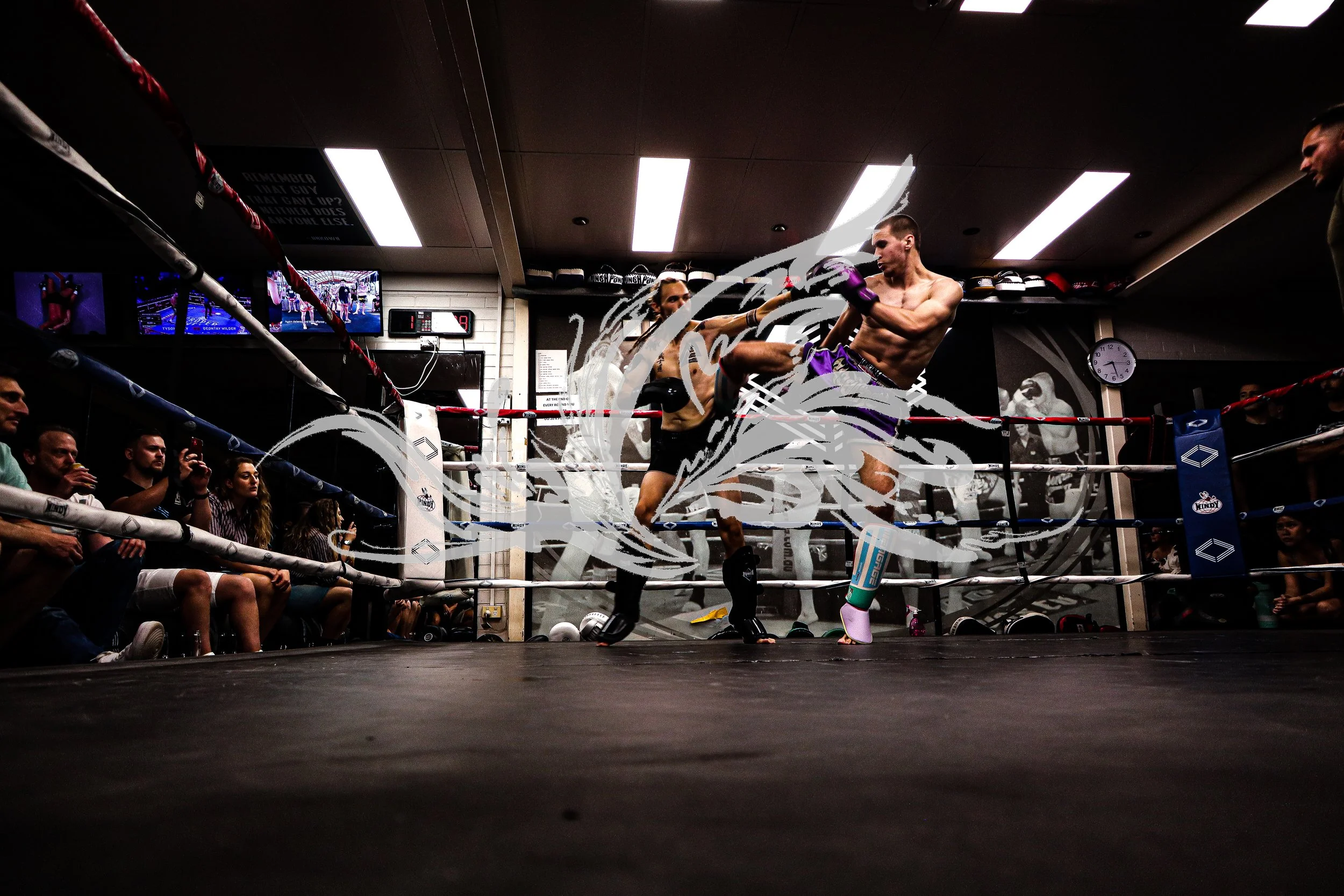 Two male fighters engaged in a kickboxing match inside a boxing ring surrounded by spectators. One fighter is delivering a kick to the other's torso, both wearing boxing gloves and shorts. Spectators are watching intently, with some taking photos. Te