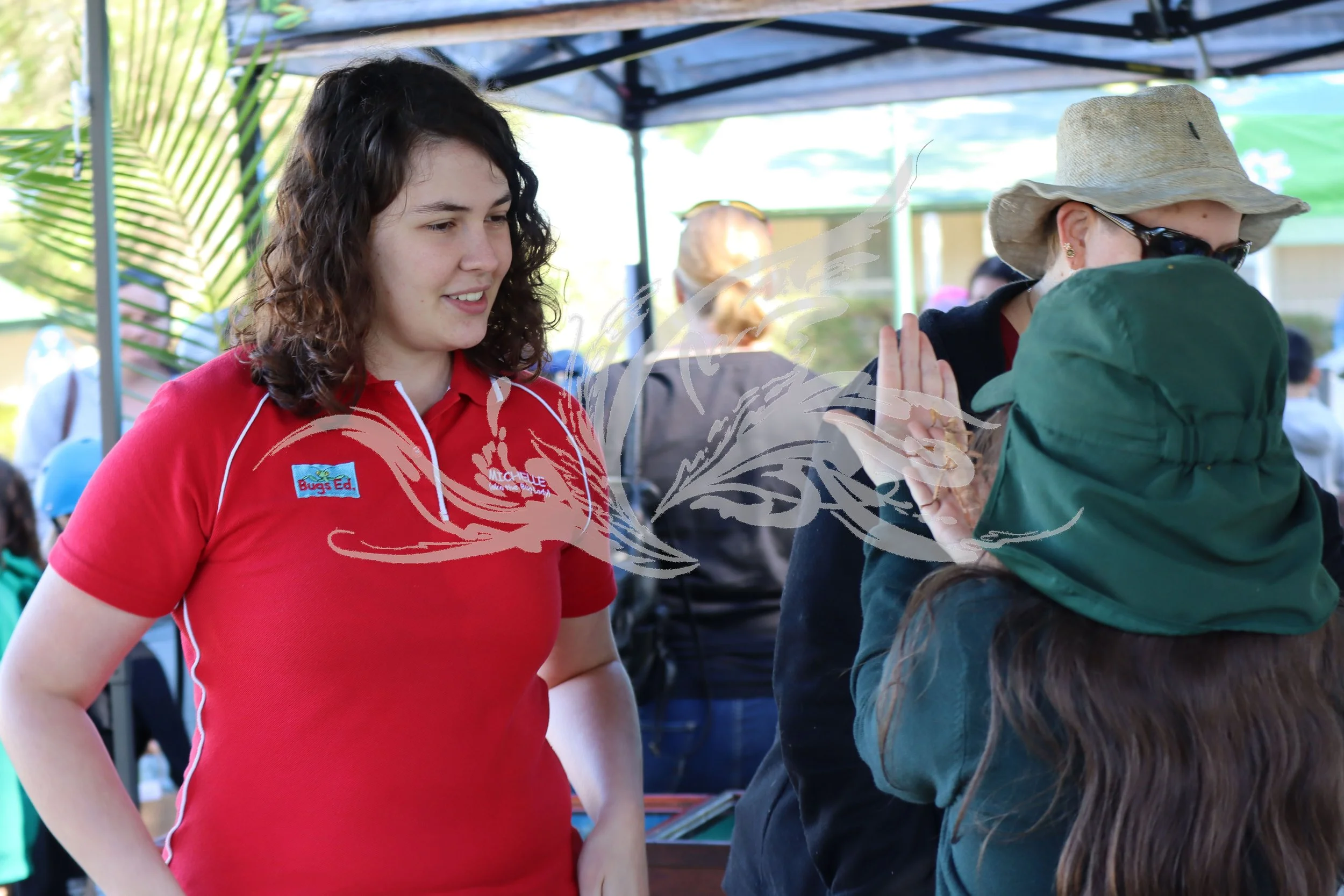 A young woman in a red polo shirt talking to another woman wearing a straw hat and sunglasses at an outdoor event under a canopy.