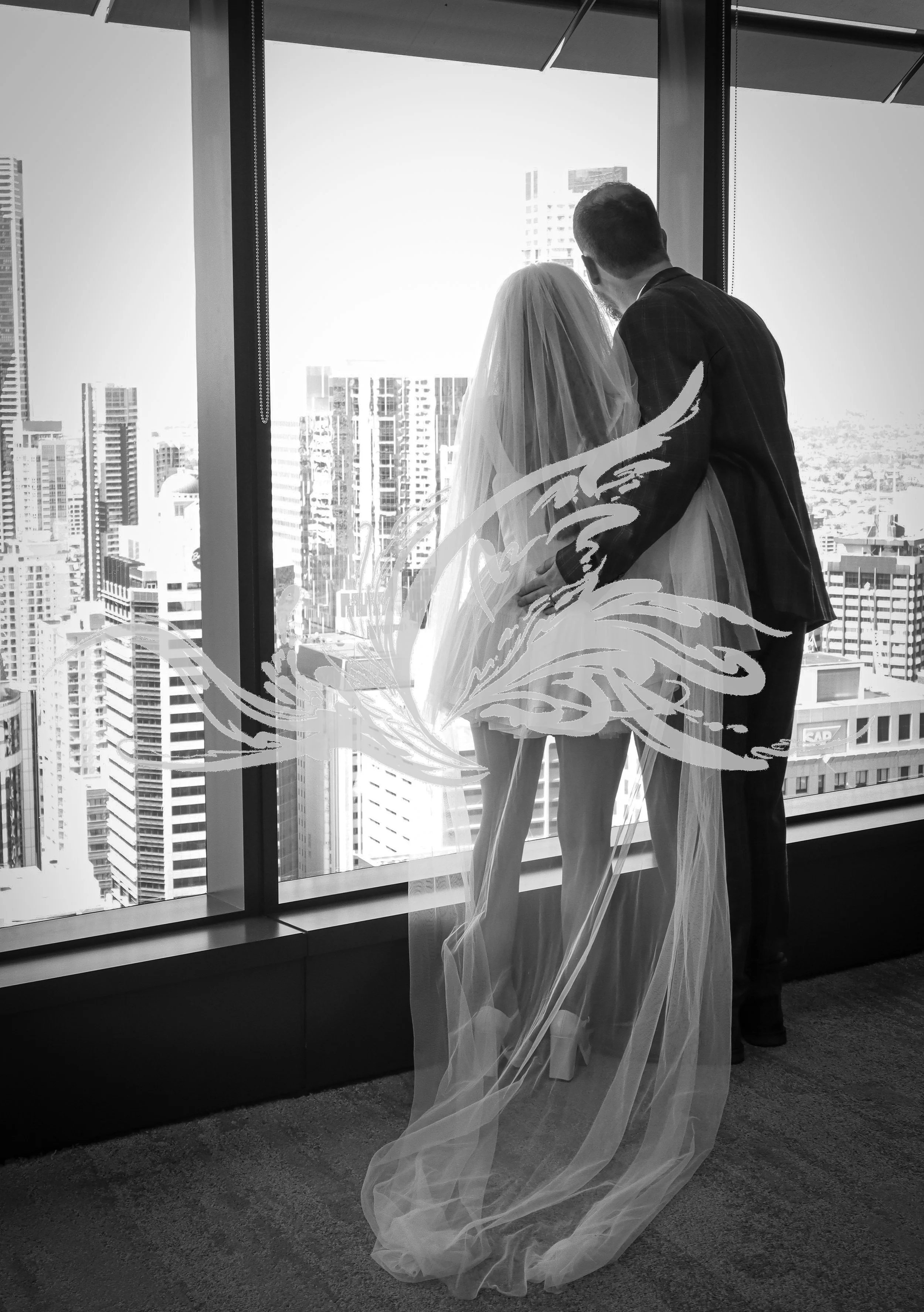 A bride and groom standing together inside a high-rise building, gazing out at a cityscape through large windows. The bride wears a wedding dress and veil, and the groom is dressed in a suit. There are tall buildings visible outside.