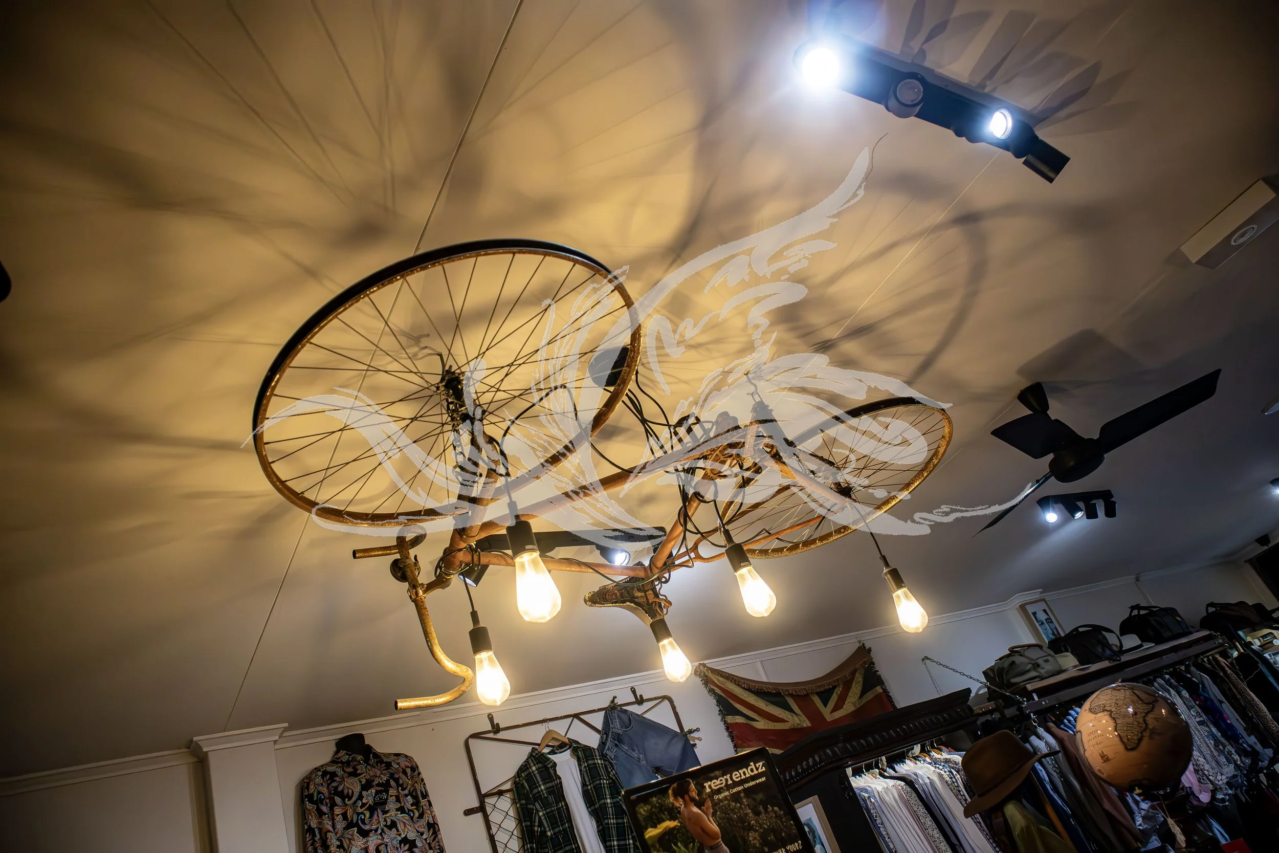 Decorative ceiling light fixture resembling bicycle wheels with exposed light bulbs, attached to a wooden ceiling with ceiling fan and store interior below.