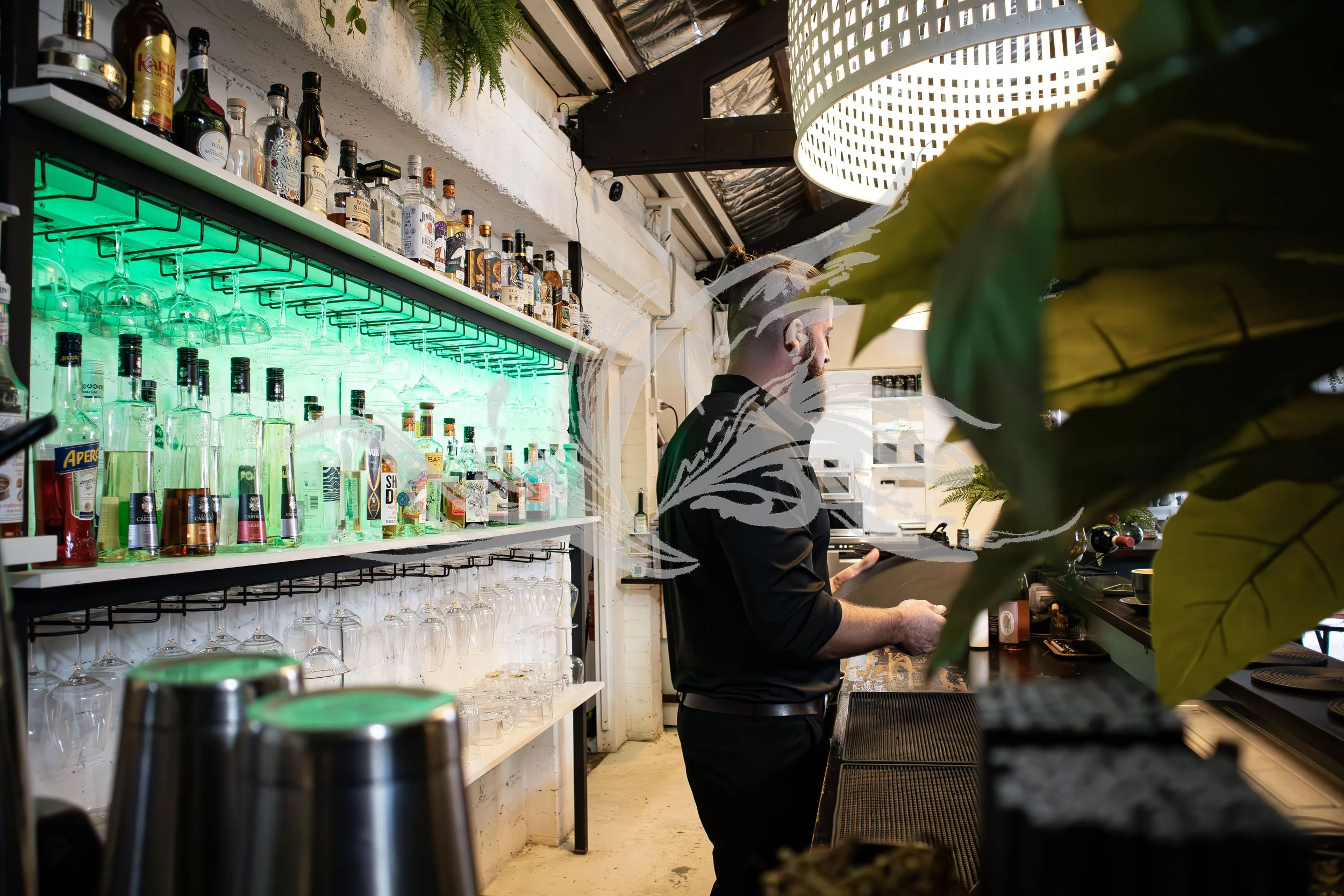 Bar interior with bottles of alcohol, wine glasses, and bar tools on shelves. A bartender in a black shirt is preparing drinks behind the counter, partially obscured by leaves in foreground. Green lighting illuminates the bottles.
