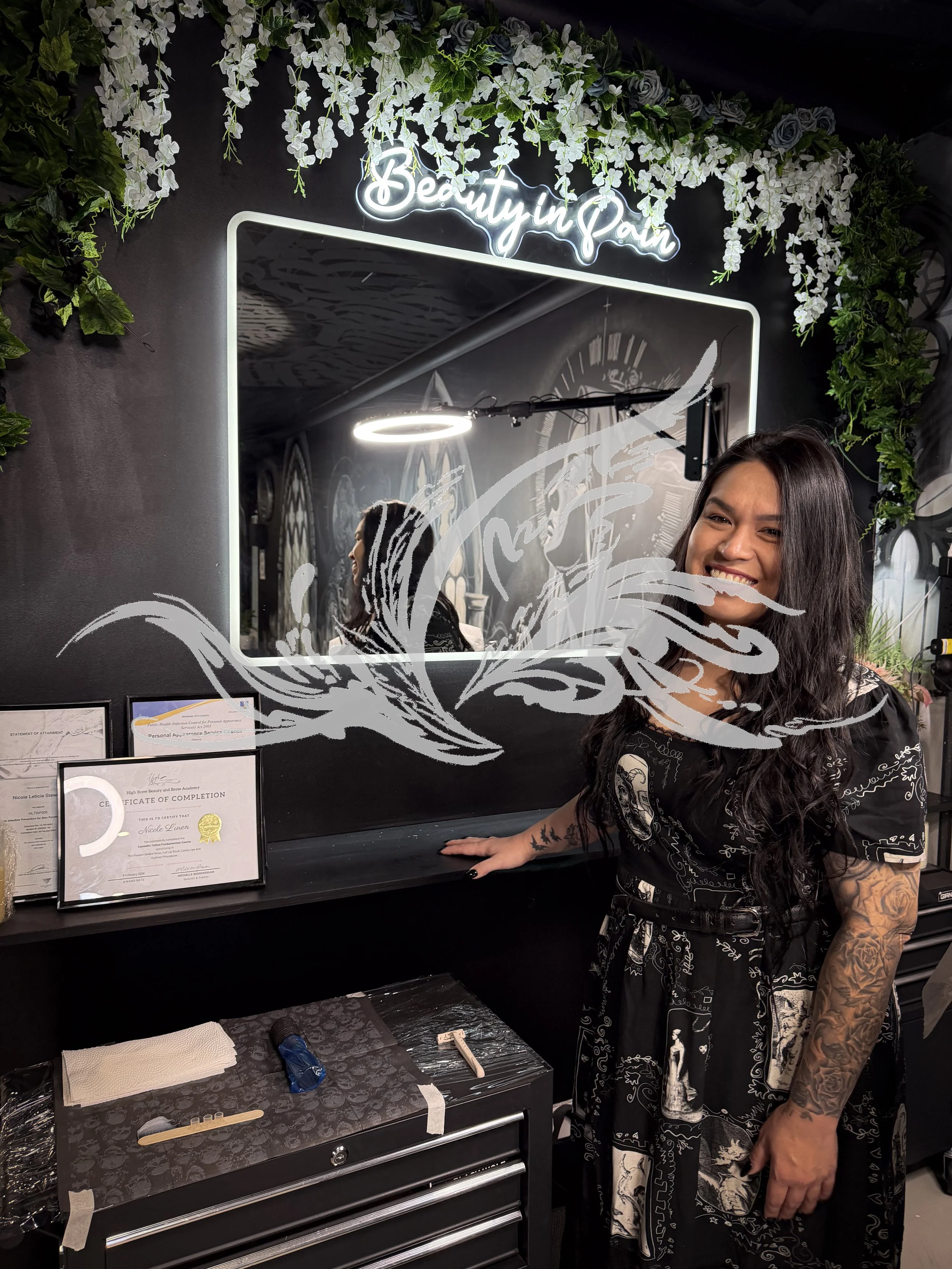 A woman with long dark hair and tattoos smiling while standing in front of a large mirror with a neon sign that reads 'Beauty in Pain' and decorative white flowers hanging above. There are certificates and a work station with tools and supplies on a 