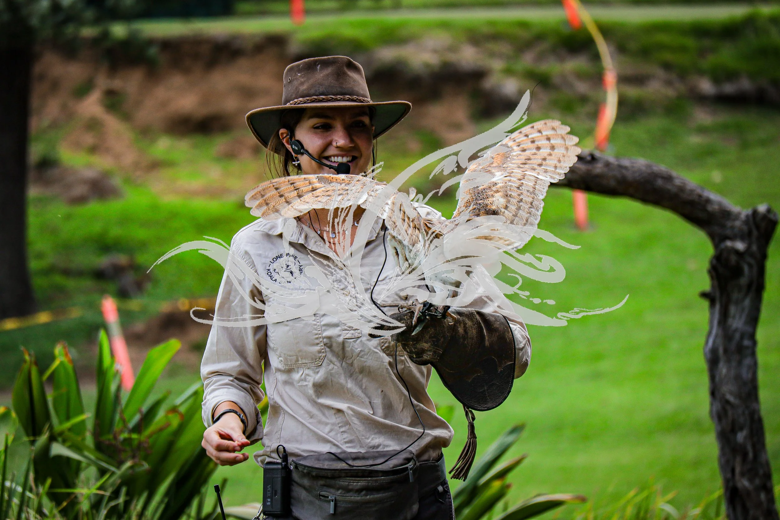 A woman outdoors wearing a wide-brimmed hat, headset, and safari-style shirt with a bird perched on her arm, surrounded by greenery.